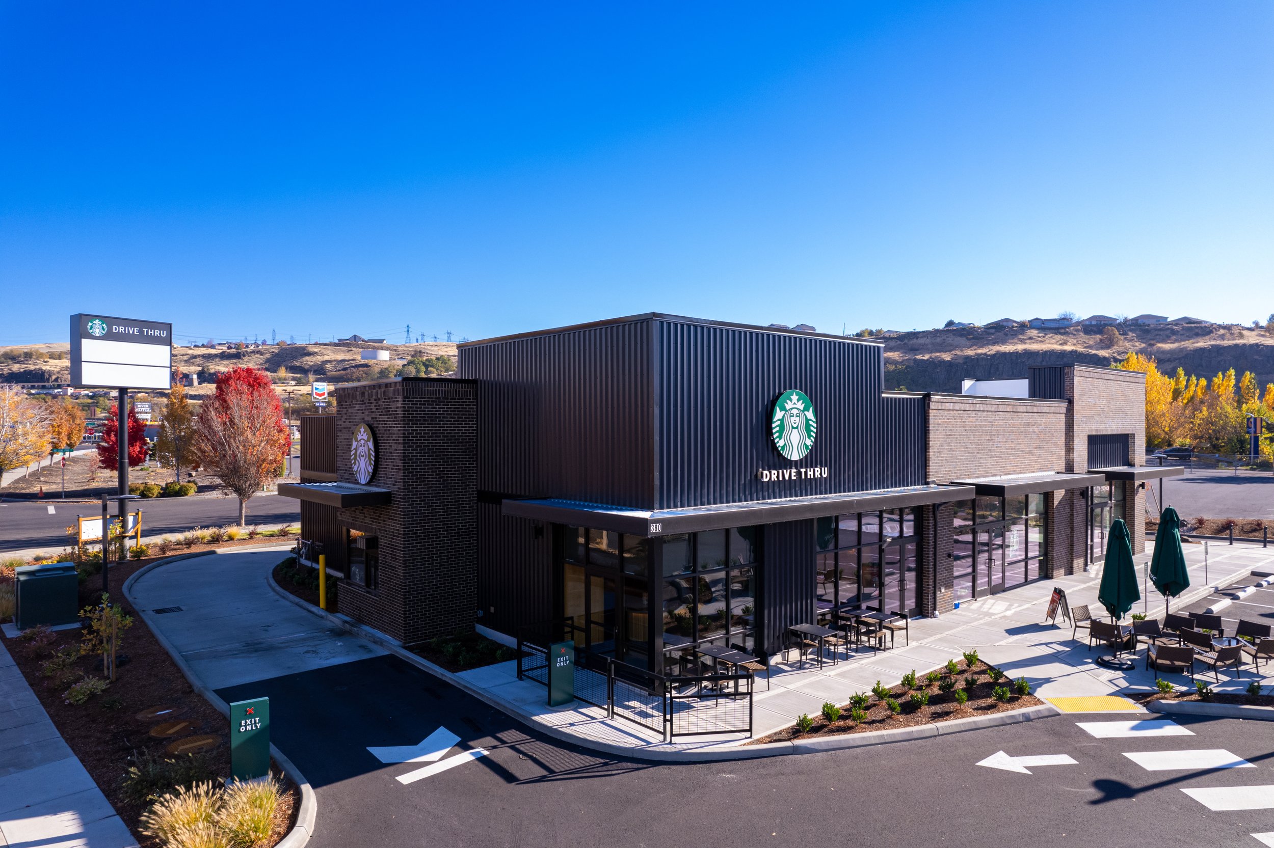 A modern Starbucks coffee shop with a drive-thru, outdoor seating with green umbrellas, and a large Starbucks logo on the black building facade, set against a clear blue sky and autumn-colored trees.
