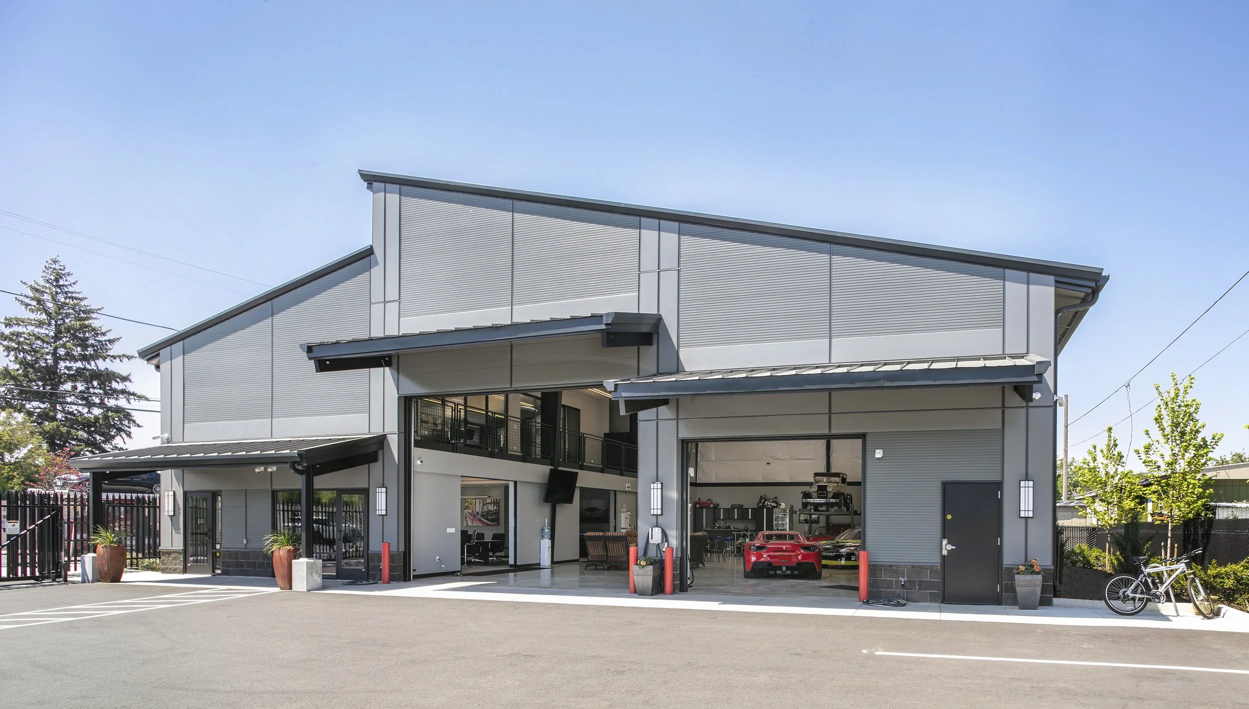 Modern multi-level building with an open garage showing cars inside, glass doors, and bicycle outside, surrounded by trees and a paved parking lot.