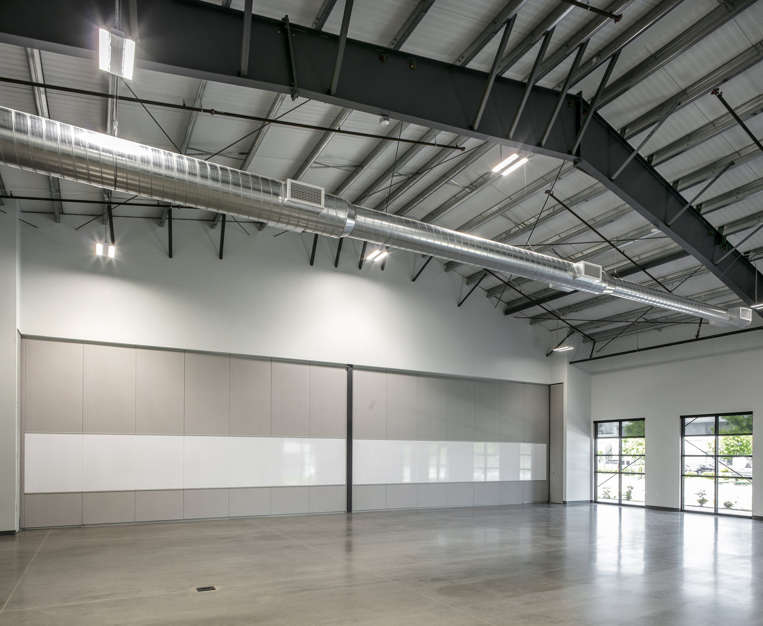 Empty industrial or commercial warehouse space with high ceilings, large windows, and metallic ducts and beams.