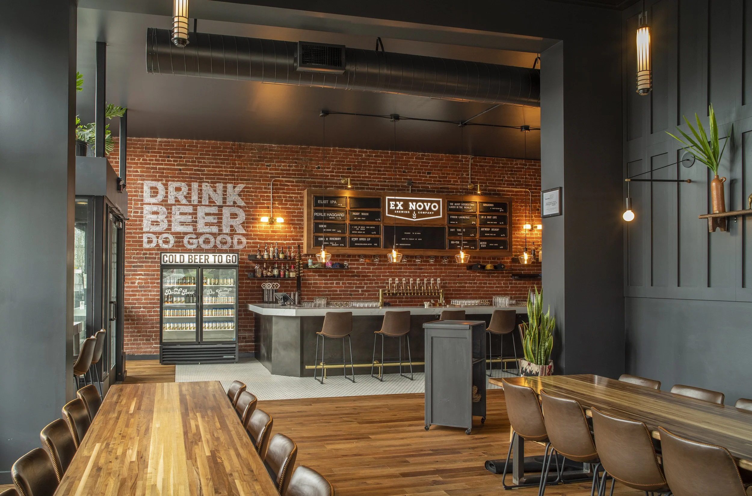Interior of a modern bar with wooden tables and chairs, brick wall with signage, and a bar counter with bottles, glassware, and a beer fridge.
