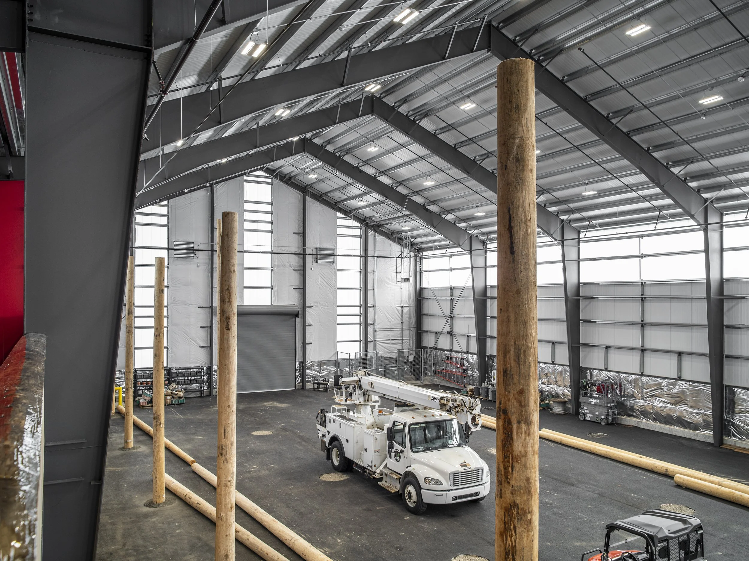Interior of a large industrial building with metal walls and ceiling, showing wooden logs and utility vehicles, including a bucket truck, with construction materials stored along the walls.