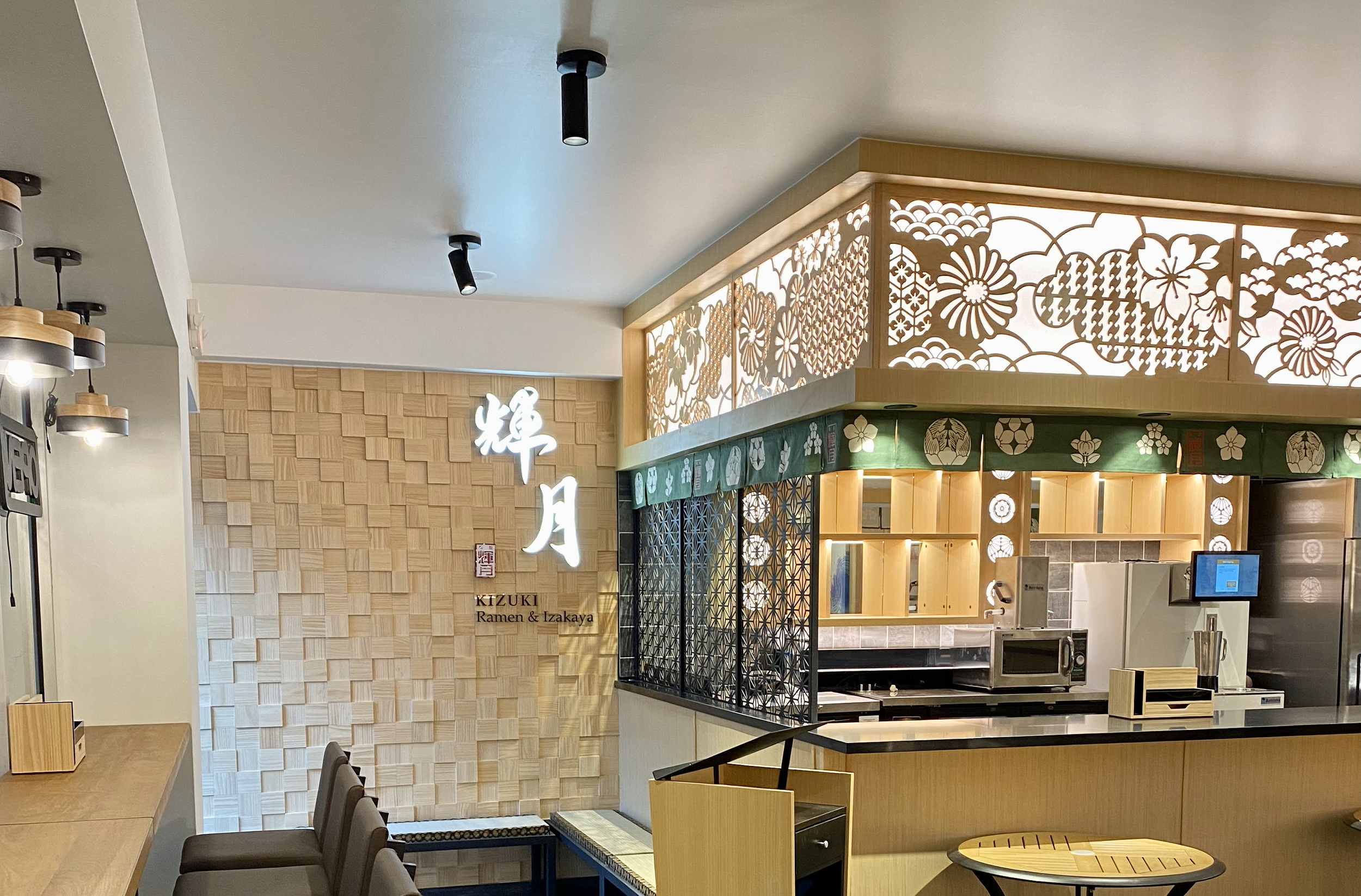 Interior of a Japanese ramen and izakaya restaurant with wooden decor, decorative lattice panels, and illuminated signage in Japanese characters.