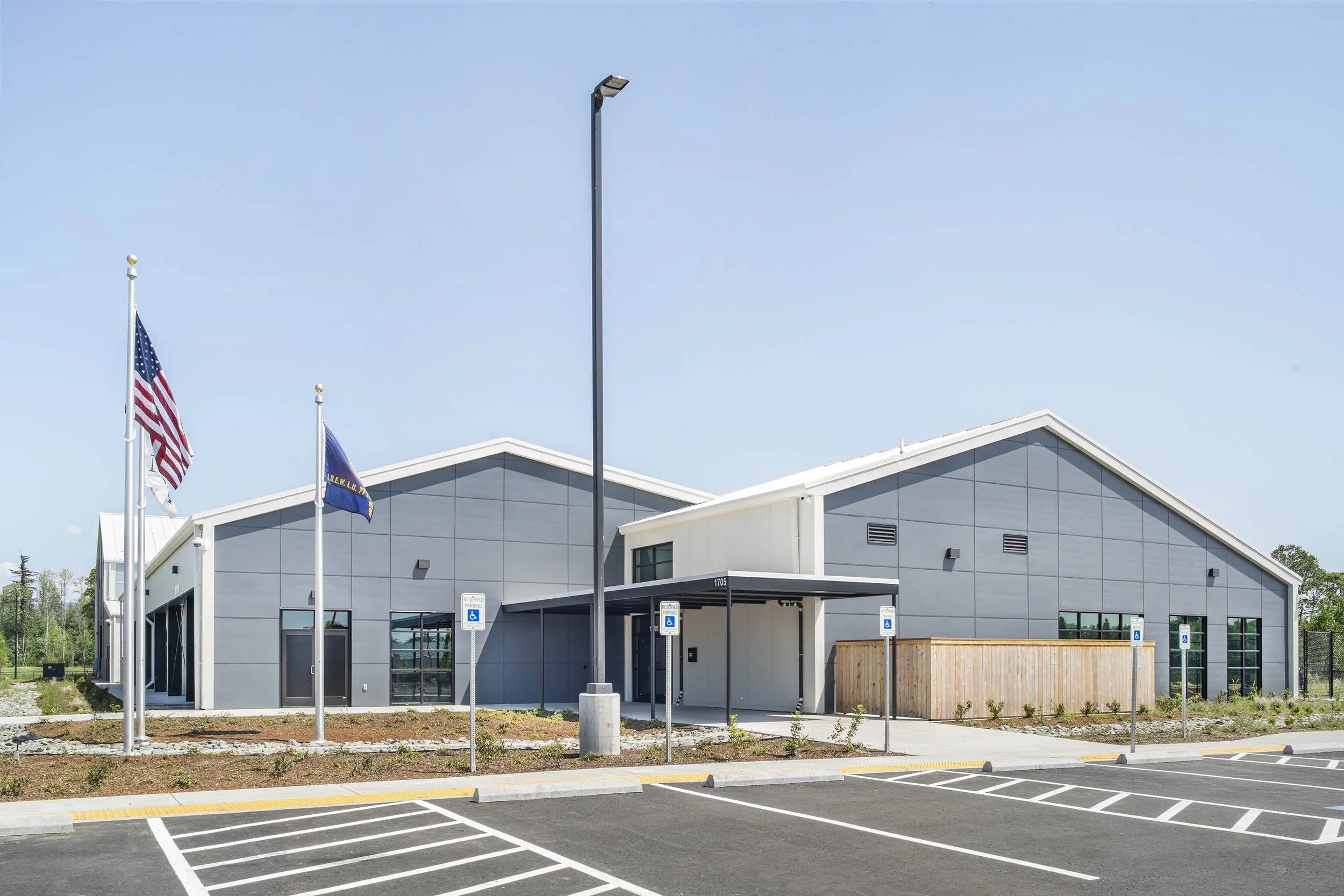 Modern building with gray panels, surrounded by a parking lot with designated handicapped parking spaces, three flags including the United States flag, and a clear blue sky.