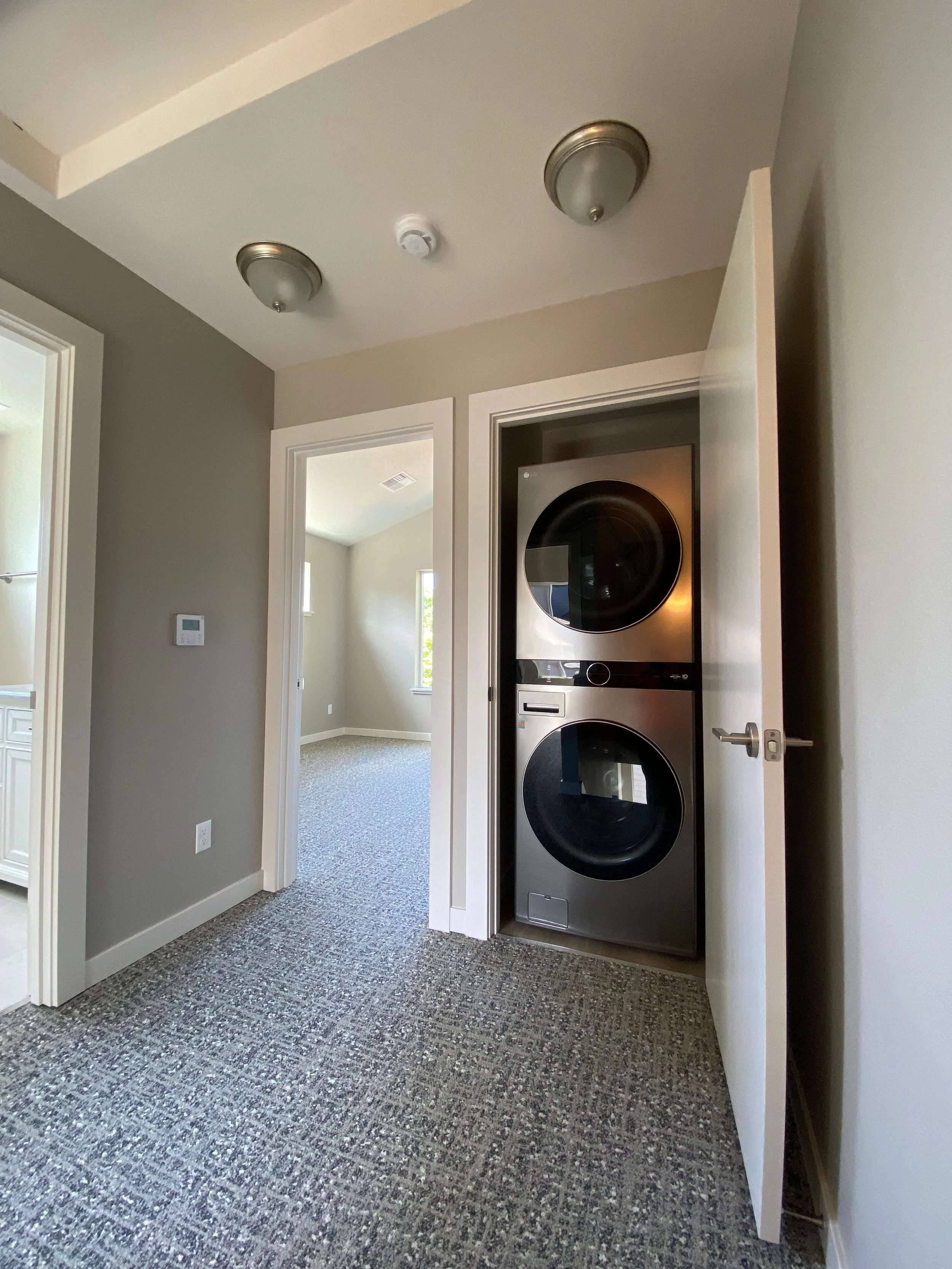 A laundry room with stacked washer and dryer inside a closet with a door propped open. There are two ceiling lights and a smoke detector on the ceiling, and a thermostat on the wall. The room has gray walls and textured carpet floors.