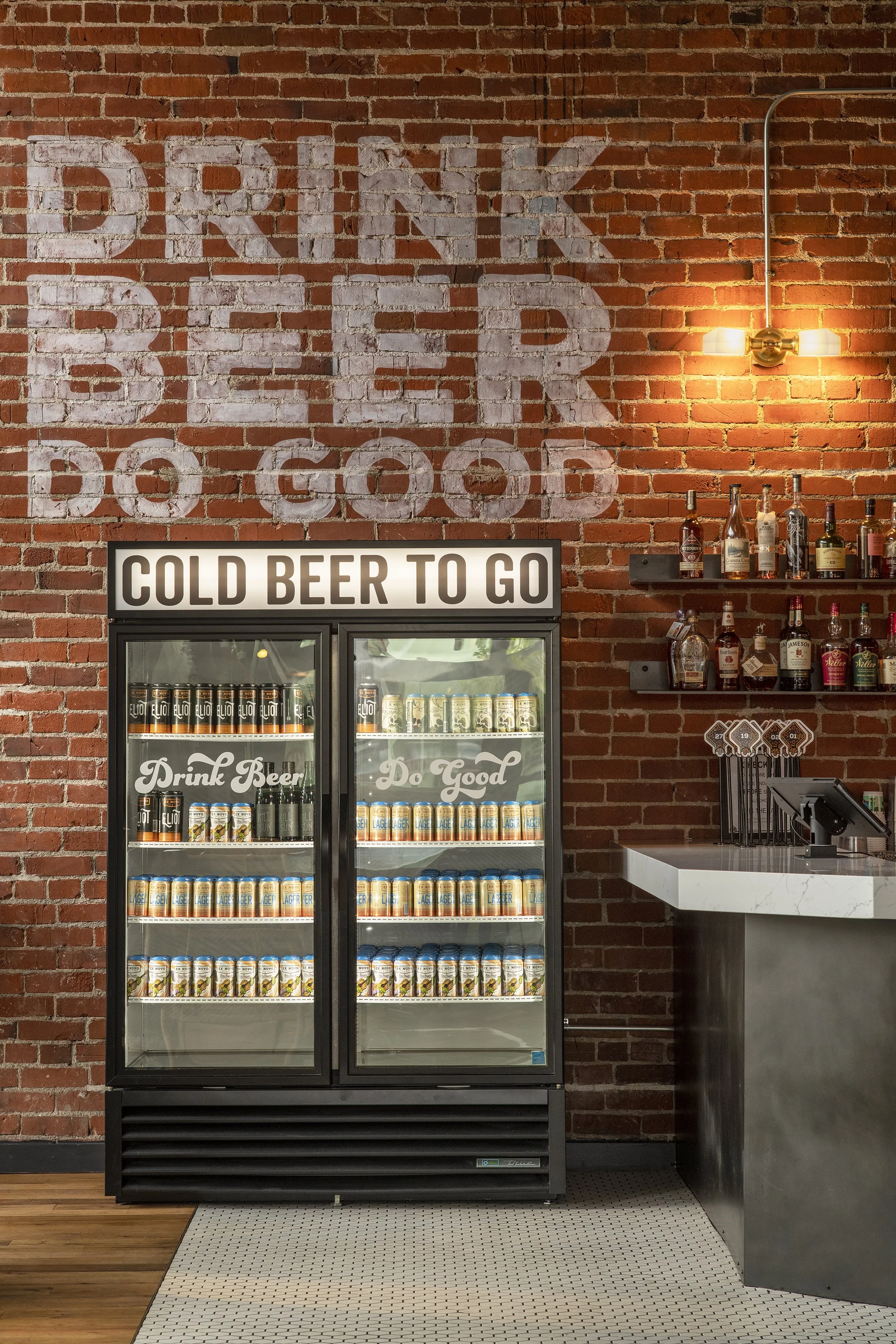 A refrigerator stocked with cans and bottles of beer, with a sign above that says 'Cold Beer To Go', and a brick wall with a painted sign reading 'Drink Beer Do Good'. To the right, there are shelves with bottles of alcohol and a bar counter.