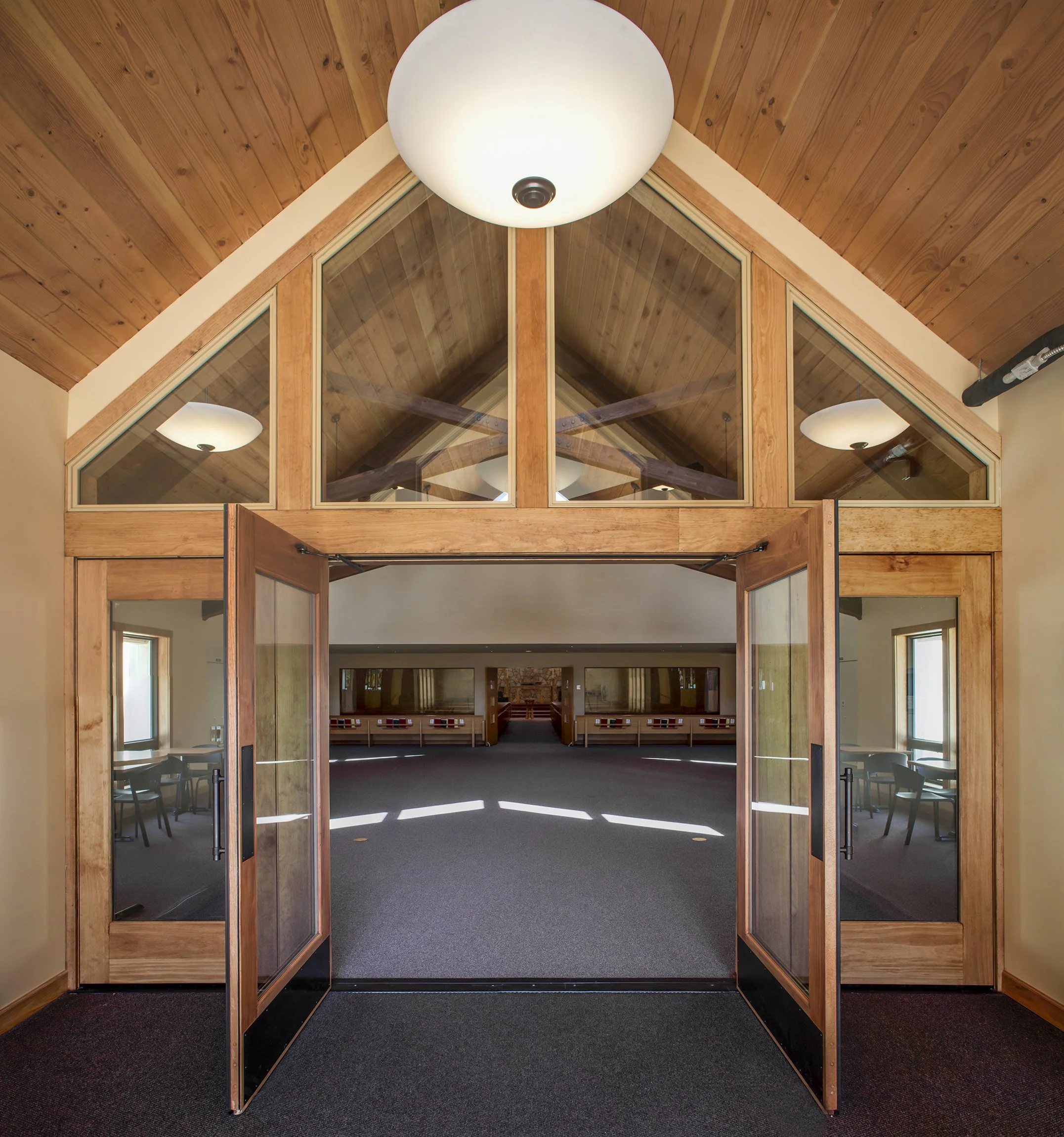 Interior view of a wooden building with double glass doors and a vaulted ceiling with wood paneling, featuring chandeliers and windows letting in natural light.