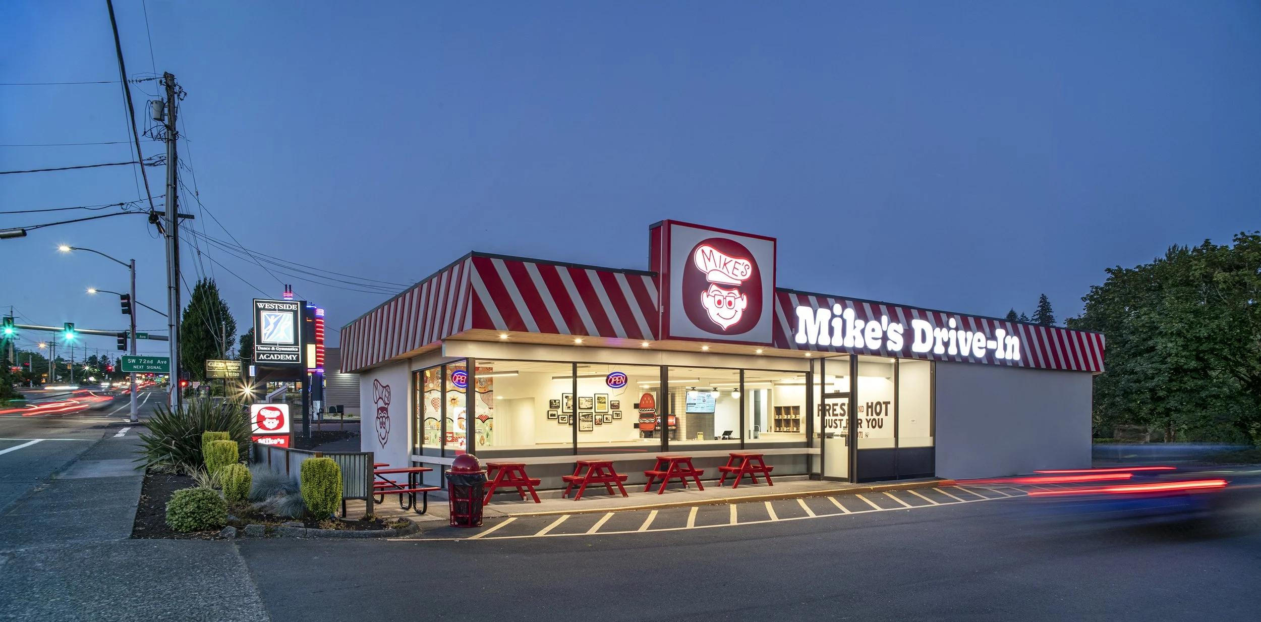 Exterior of Mike's Drive-In restaurant during evening, with bright signs, red picnic tables outside, and a large lit sign with a cartoon face wearing a burger hat.