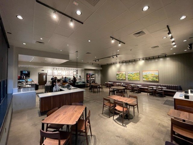 Empty cafe or restaurant interior with wooden tables and chairs, a counter area in the background, and three landscape paintings on the wall.