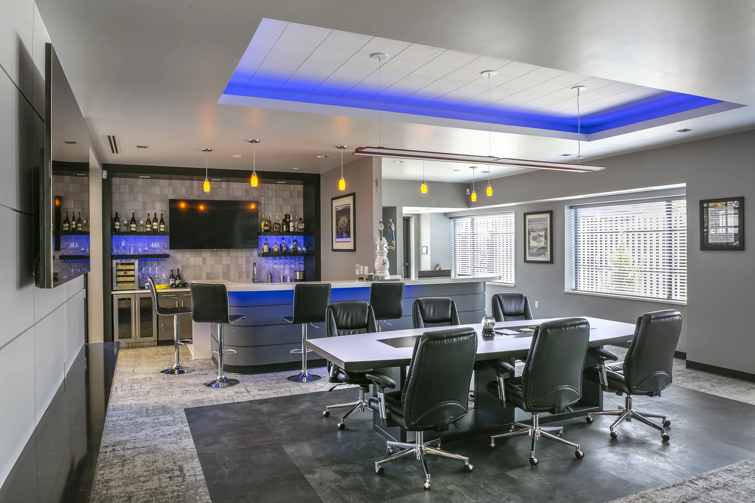 Modern conference room with a long table and black leather chairs, a bar area with a blue-lit counter, a TV on the wall, large windows with blinds, and artwork on the walls.
