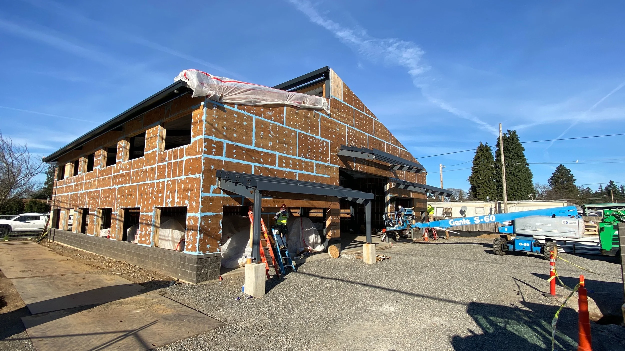 Construction site of a two-story building with unfinished brick walls, construction workers, scaffolding, and a blue Genie S-60 telescopic boom lift under a partly cloudy sky.