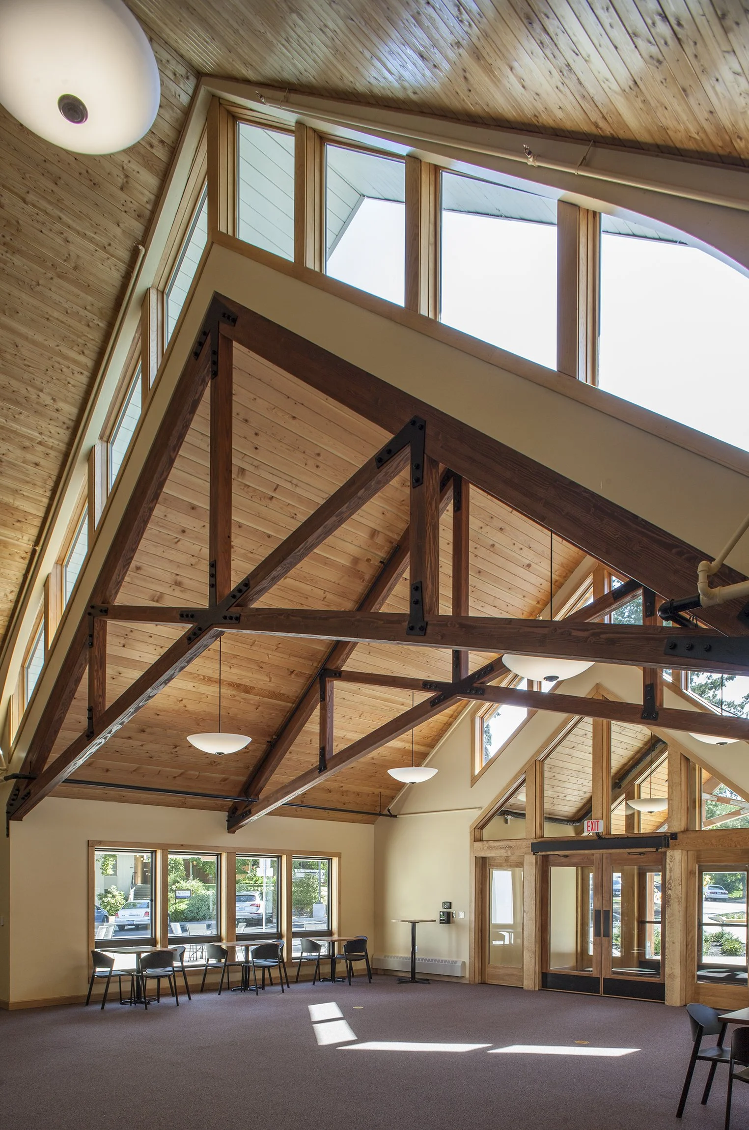 Interior of a large, bright room with wooden beams, high ceilings, windows, and a row of chairs along the wall.