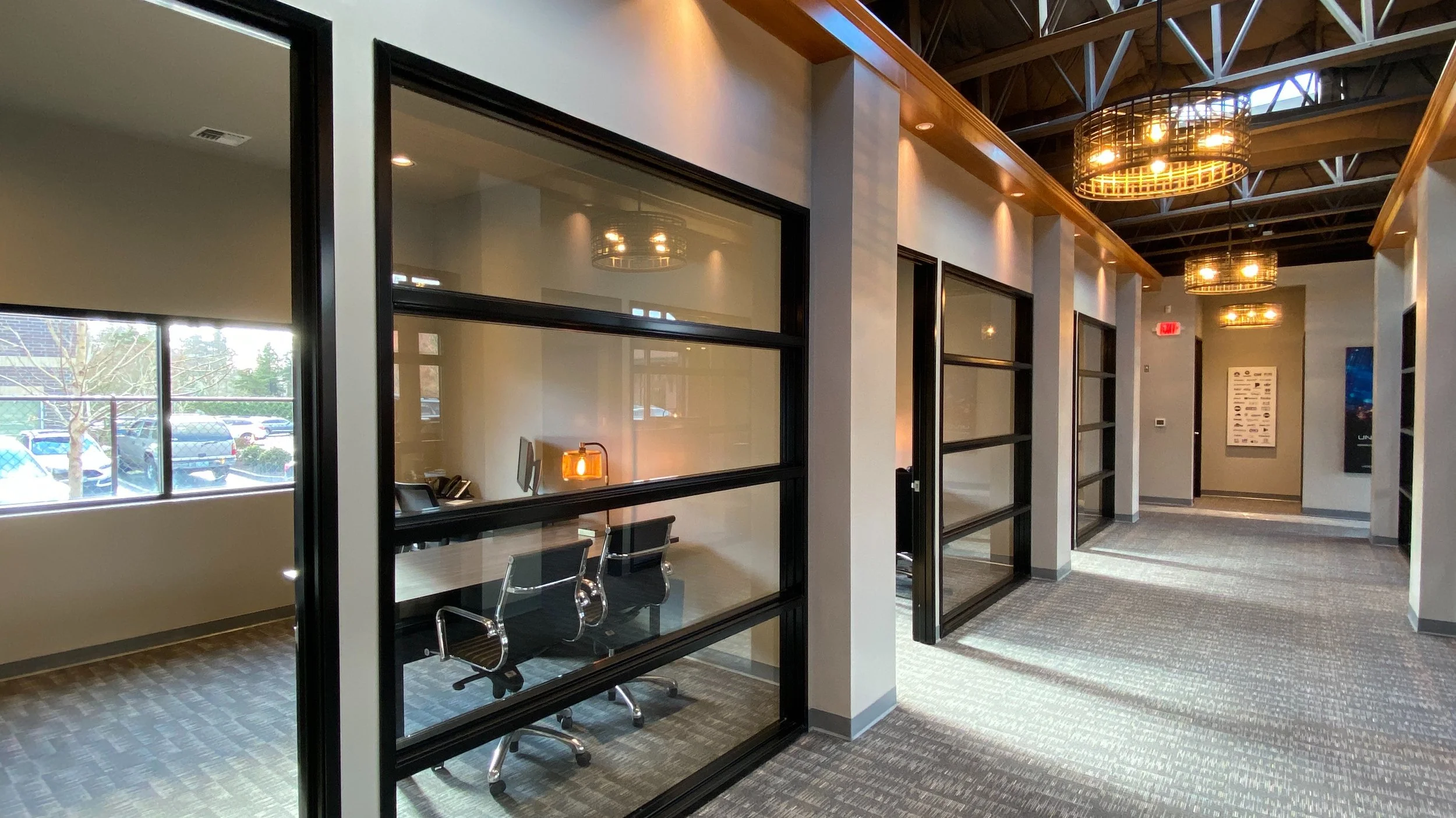 A modern office hallway with glass office walls, a conference room with a desk and chairs, and large windows letting in natural light. The hallway has hanging circular light fixtures and carpeted flooring.
