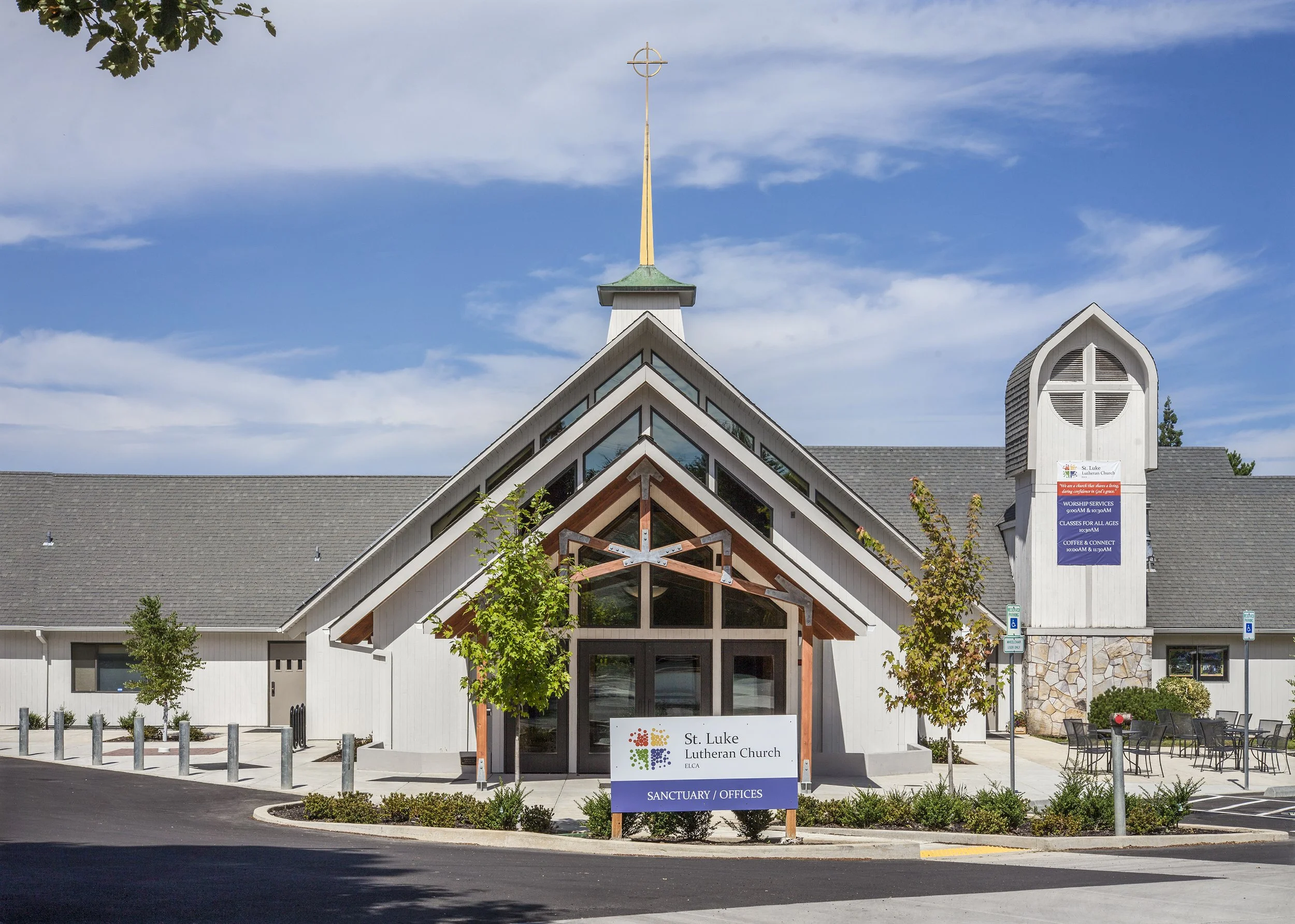 Front view of St. Luke Lutheran Church with a modern architectural design, a sign indicating the sanctuary and offices, a cross on the building, and trees in the foreground under a partly cloudy sky.