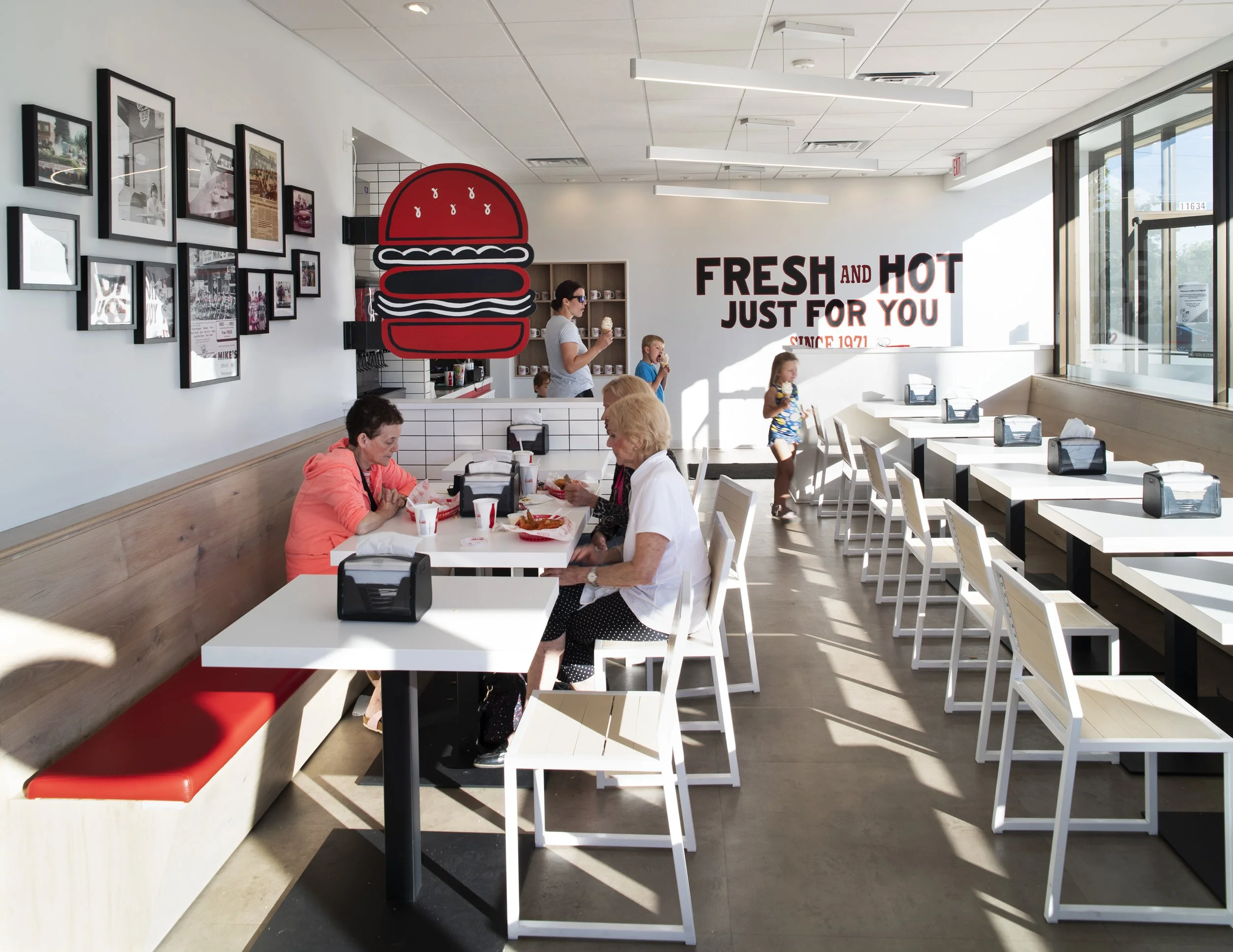 Interior of a modern fast food restaurant with white tables and chairs, some customers eating, a large red hamburger sign on the wall, and a bold sign that reads "FRESH AND HOT JUST FOR YOU SINCE 1971."