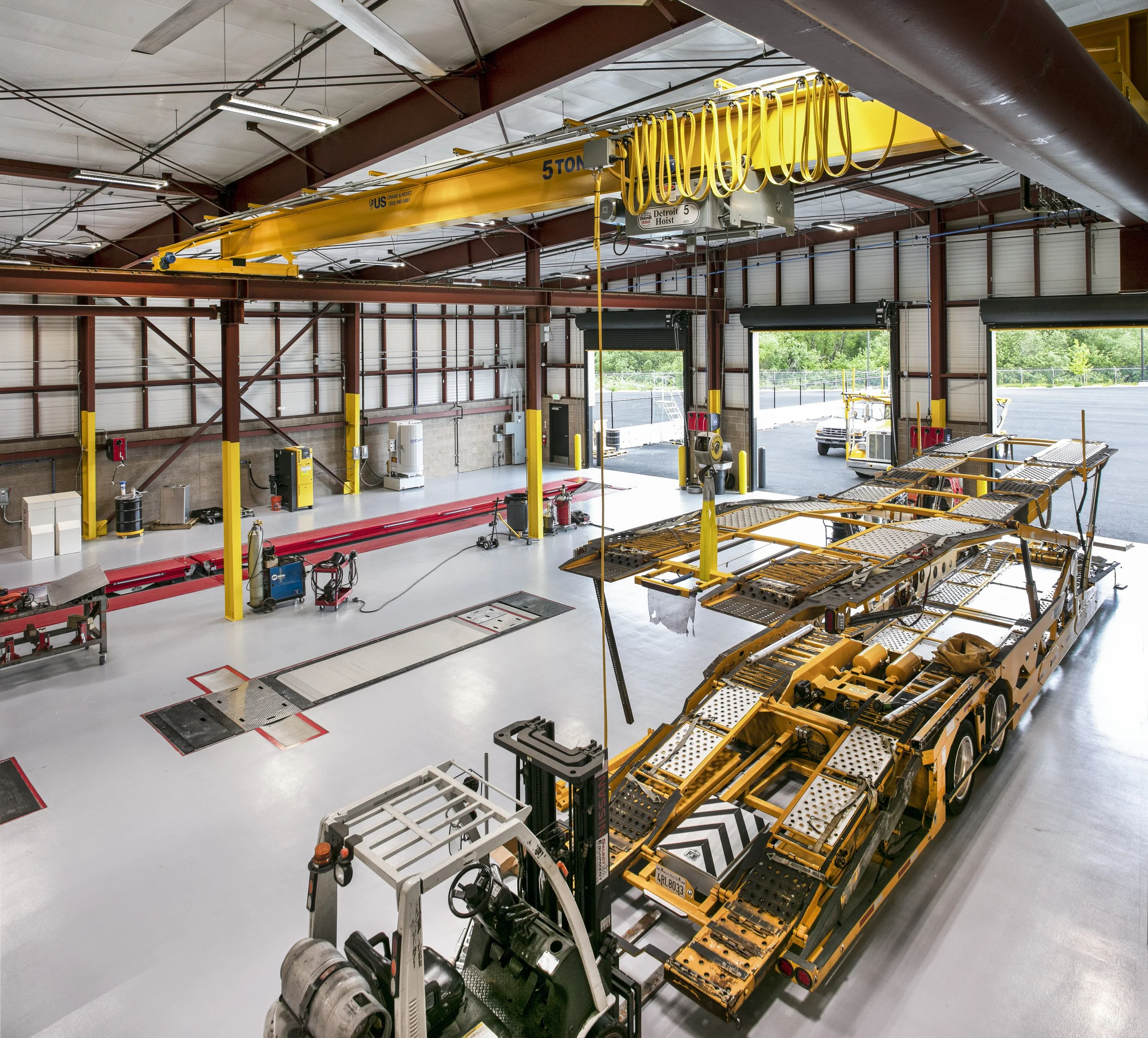 A spacious vehicle repair or maintenance garage with a large yellow lifting crane overhead, a yellow transport trailer, various tools, equipment, and opened garage doors revealing an outdoor parking lot.