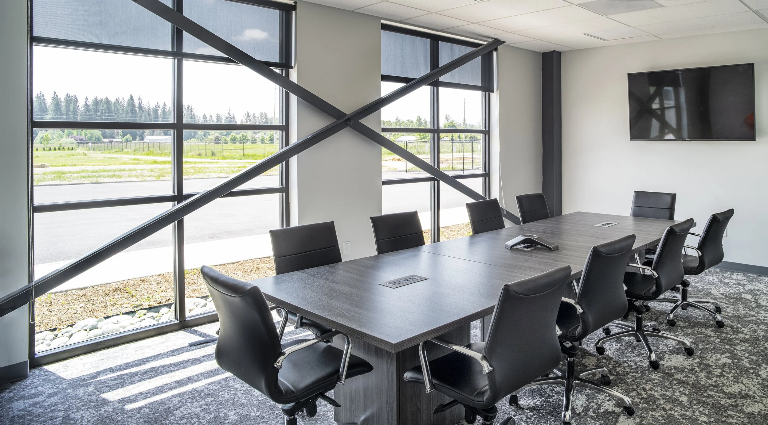 Empty modern conference room with a large table, black office chairs, large windows showing an outdoor landscape, and a wall-mounted TV.