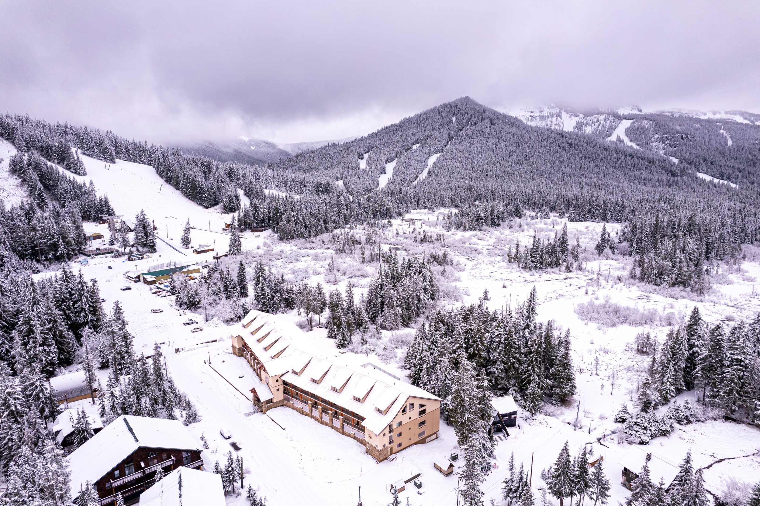 Snow-covered buildings and dense forests in a mountainous landscape during winter, cloudy sky.