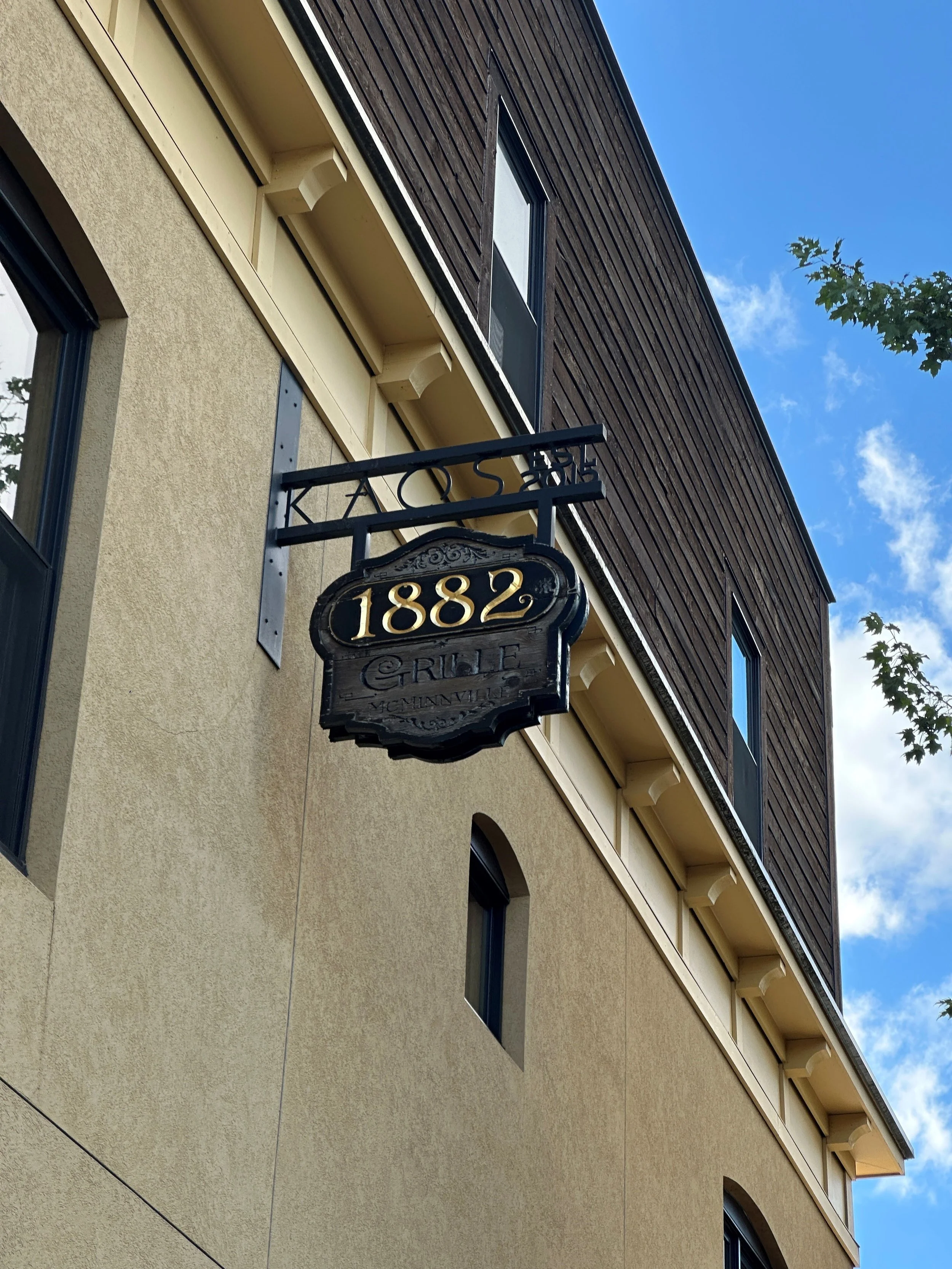 A building with a hanging sign displaying the number 1882, indicating the address, and the words 'Gaule' and 'McNuttville' below it, with a blue sky and some clouds in the background.