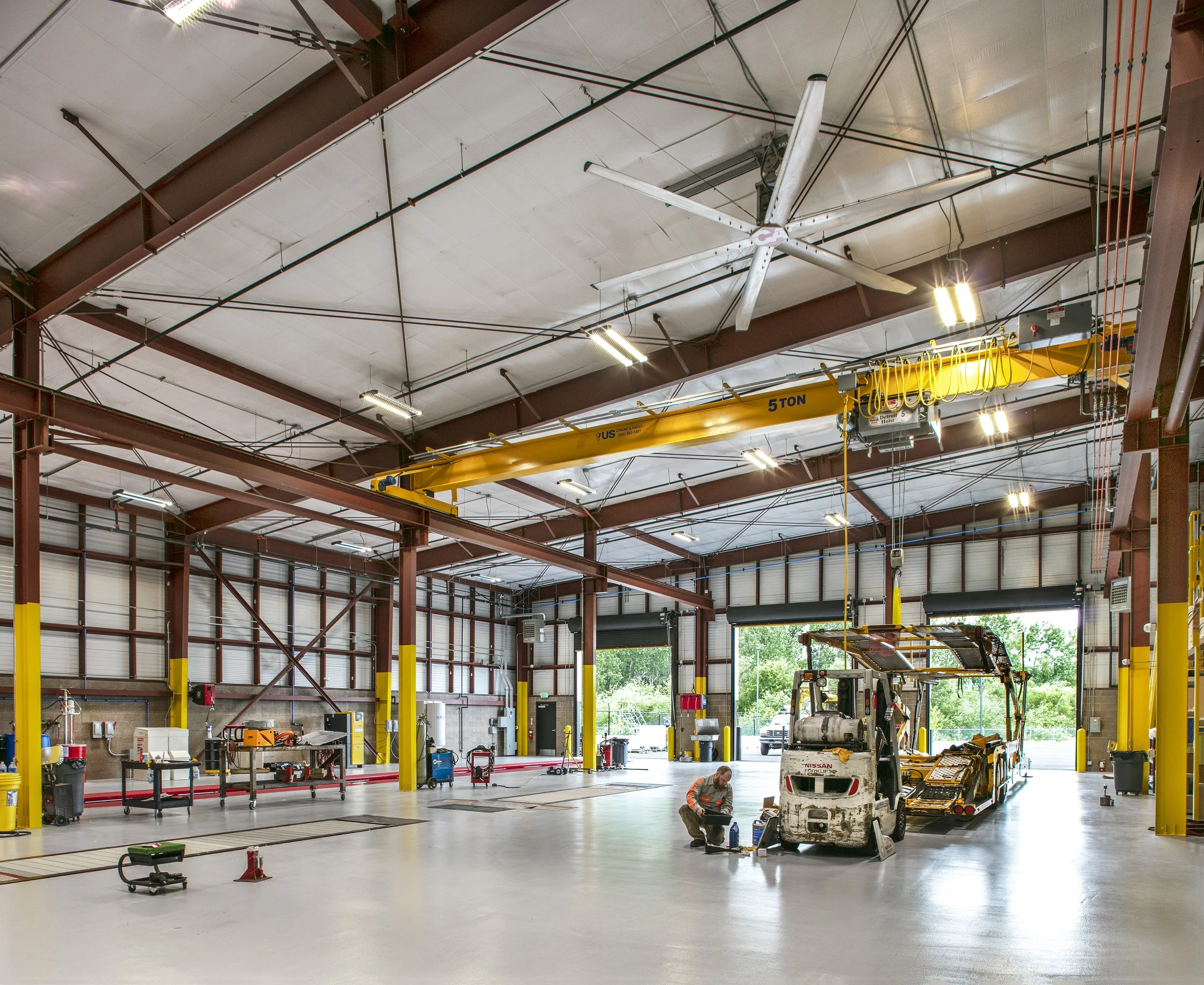 Inside an industrial warehouse, a worker repairs a golf cart with a vehicle lift and crane overhead, surrounded by tools and equipment.