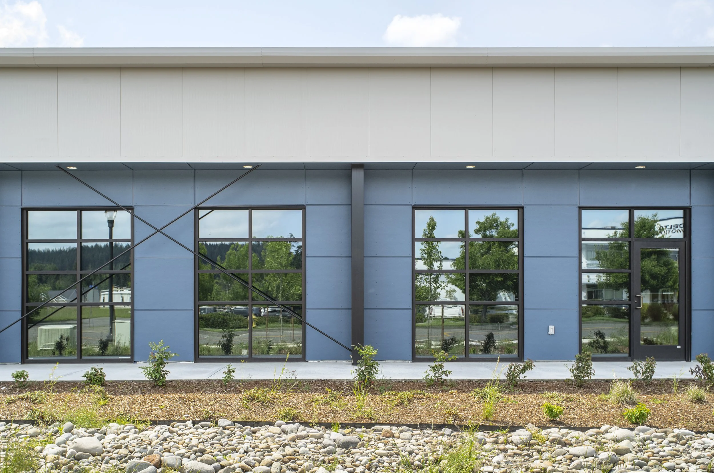 Front view of a modern commercial building with blue exterior walls, large reflective windows, and small landscaping with plants and stones in front.
