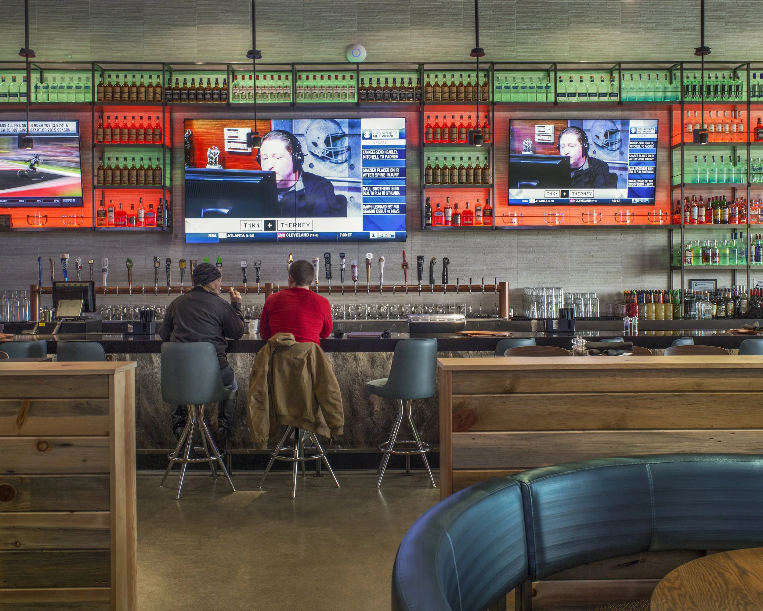 A bar scene with two people sitting at the counter, facing televisions displaying sports coverage, with shelves of liquor bottles and beer taps in the background.
