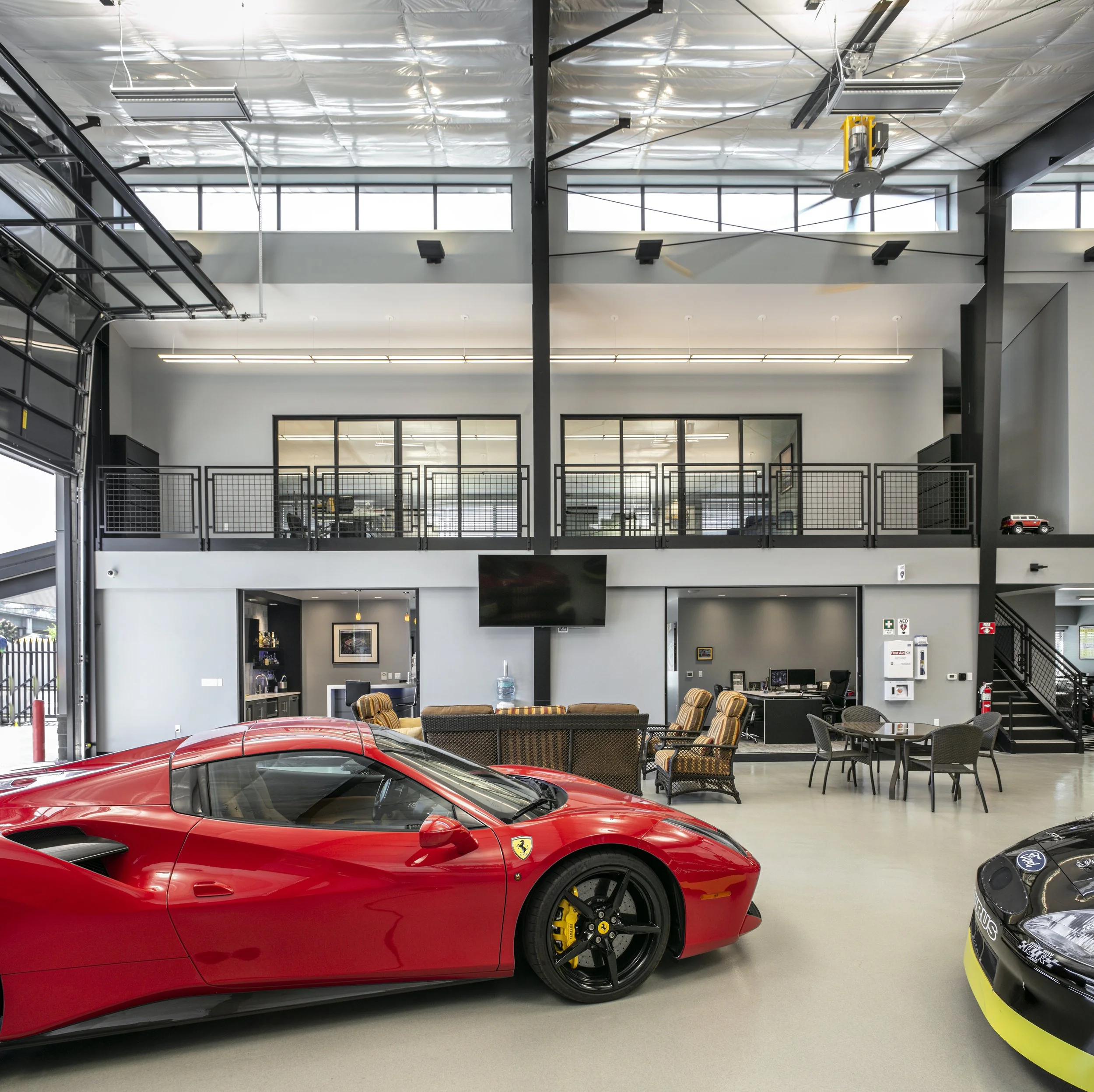 Inside a car dealership with a red Ferrari sports car in the foreground, other cars, seating area, office spaces, and a mezzanine level.