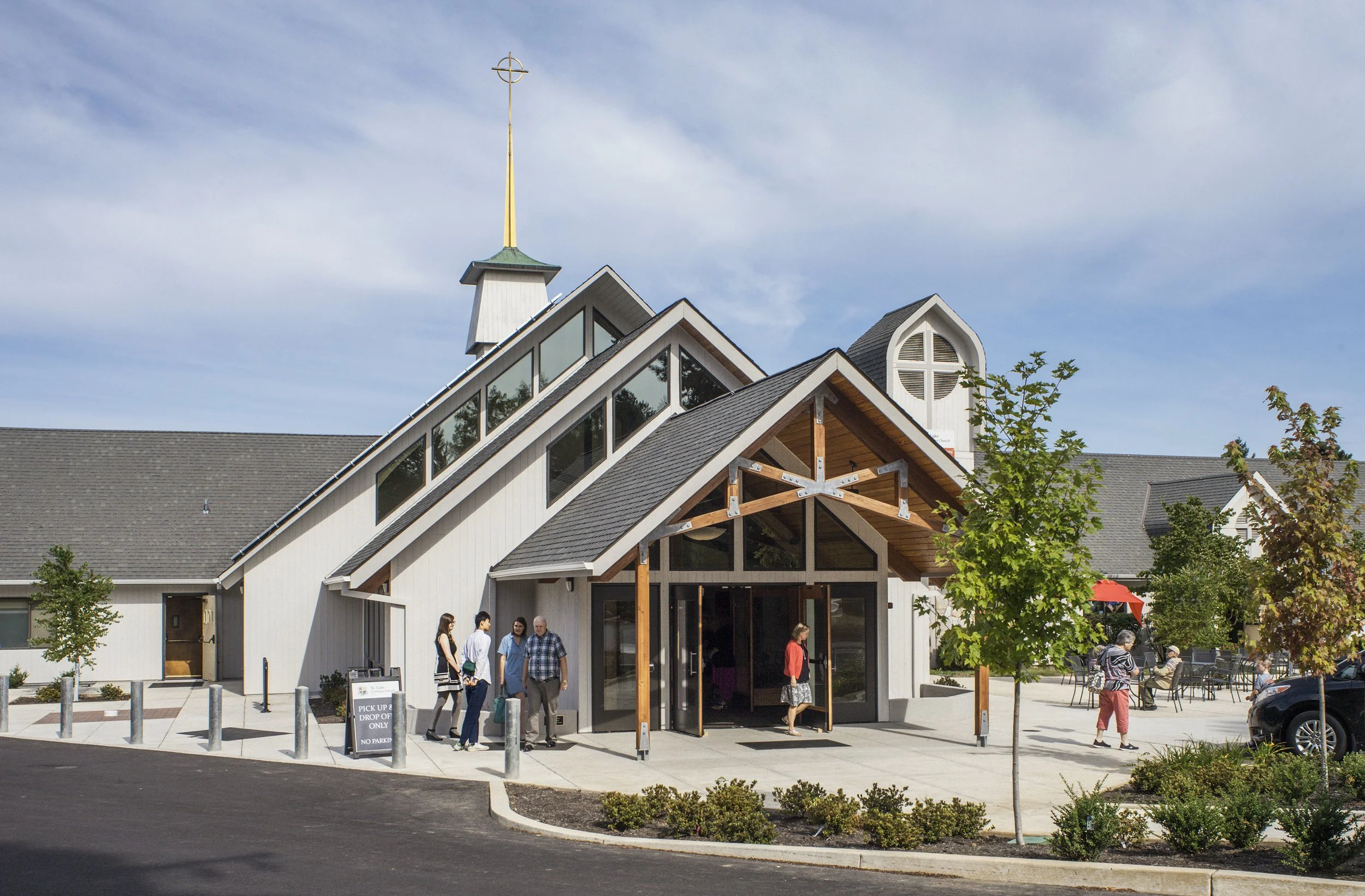 A modern church with large windows and a steep roof, people standing outside on a sunny day, some walking, some seated under a red umbrella, trees and a parking area in the background.