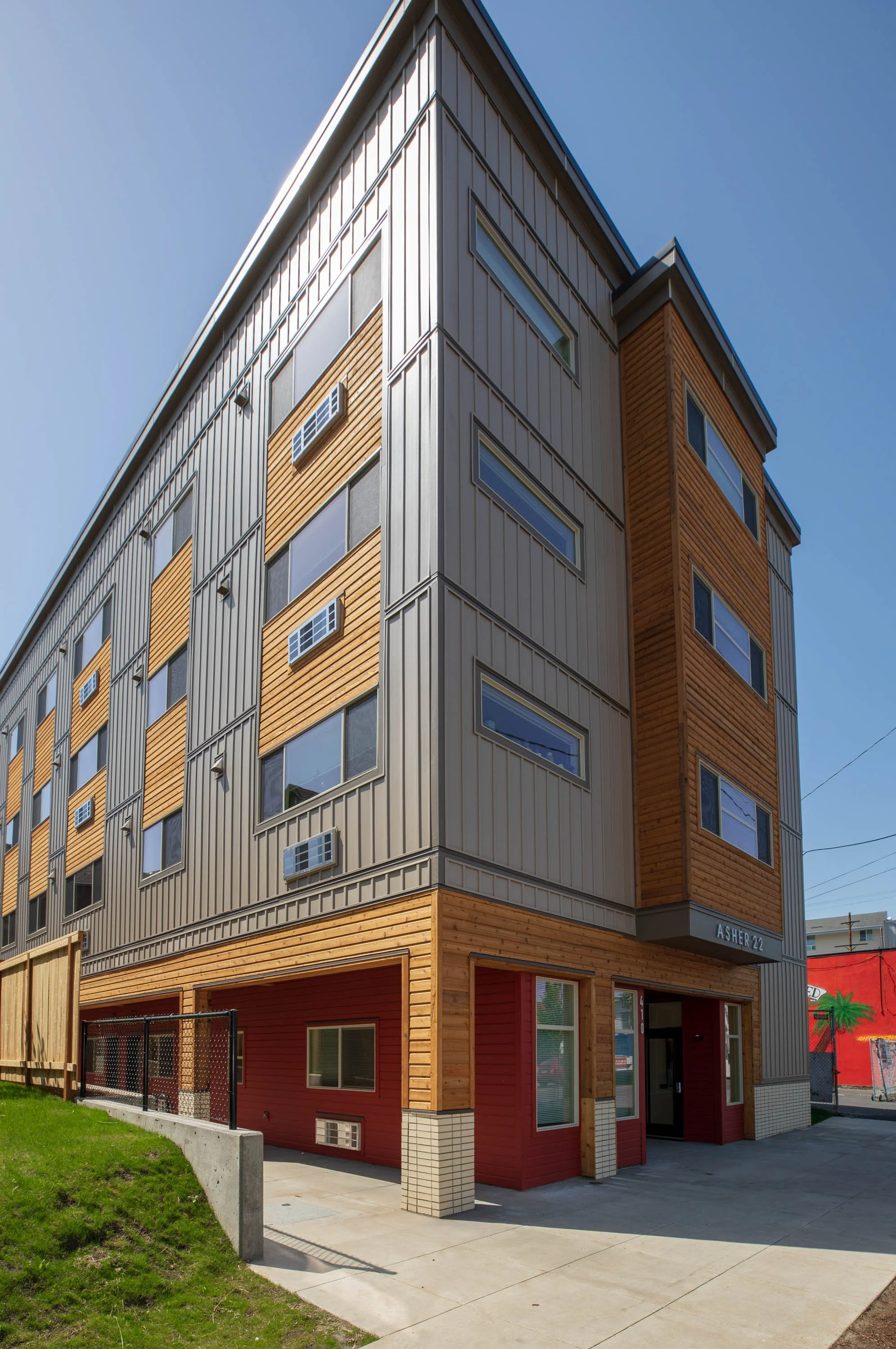 A multi-story residential building with a combination of beige, gray, and wood-paneled exterior, featuring several windows and air conditioning units, with a sidewalk, grass, and a fence in the foreground.