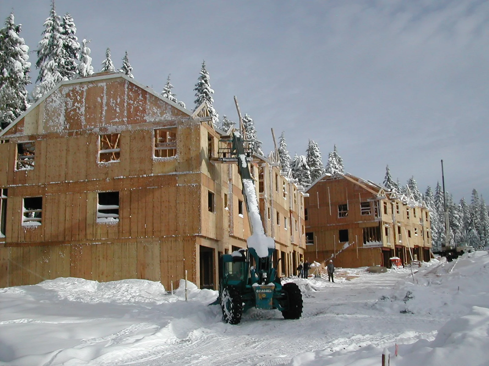 Construction site with partially built wooden houses in a snowy landscape, with a snow-covered crane and workers present, surrounded by snow-covered trees.