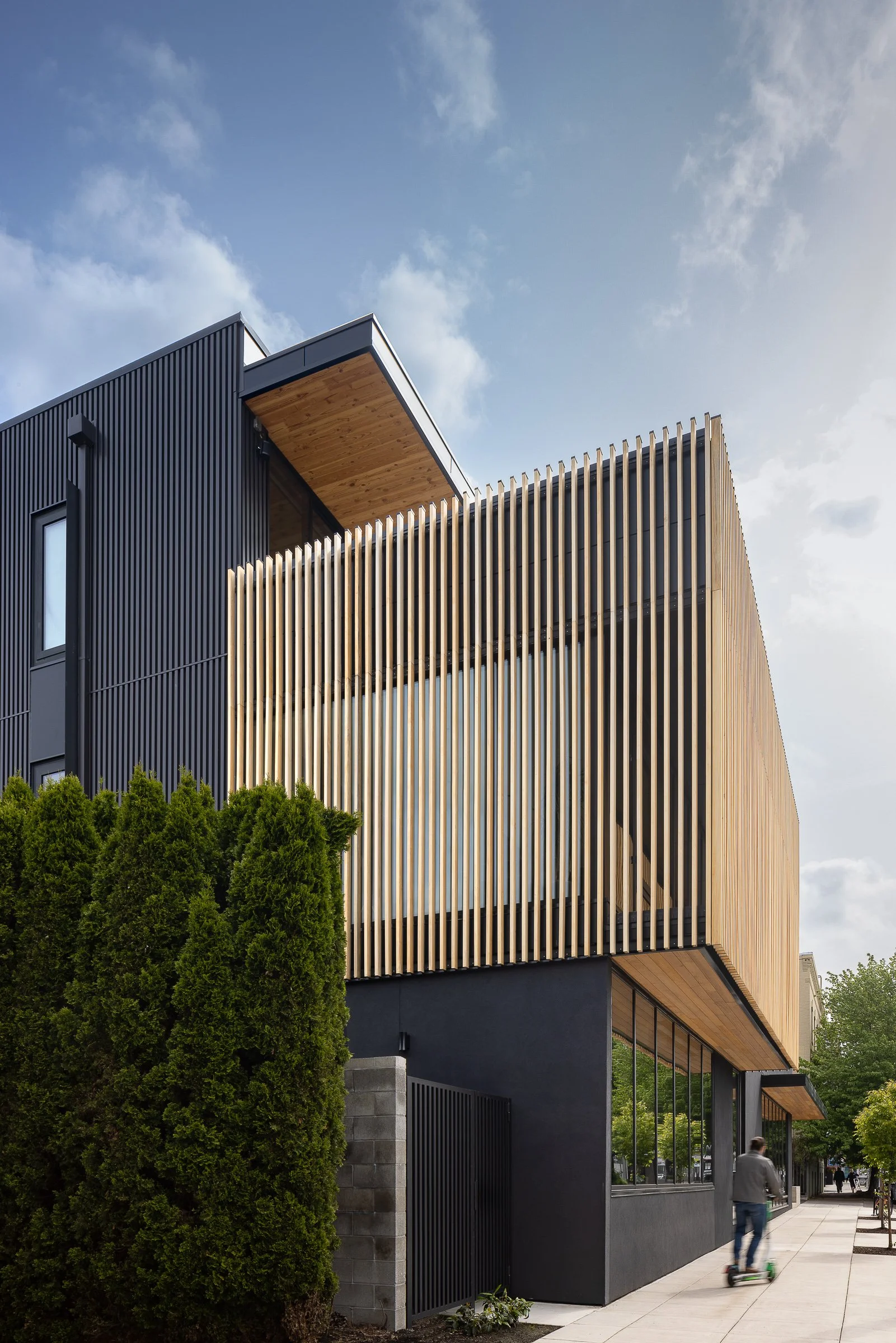 Modern building with black and wooden exterior, featuring vertical wooden slats and large windows, on a city sidewalk with a person riding an electric scooter and trees lining the street.