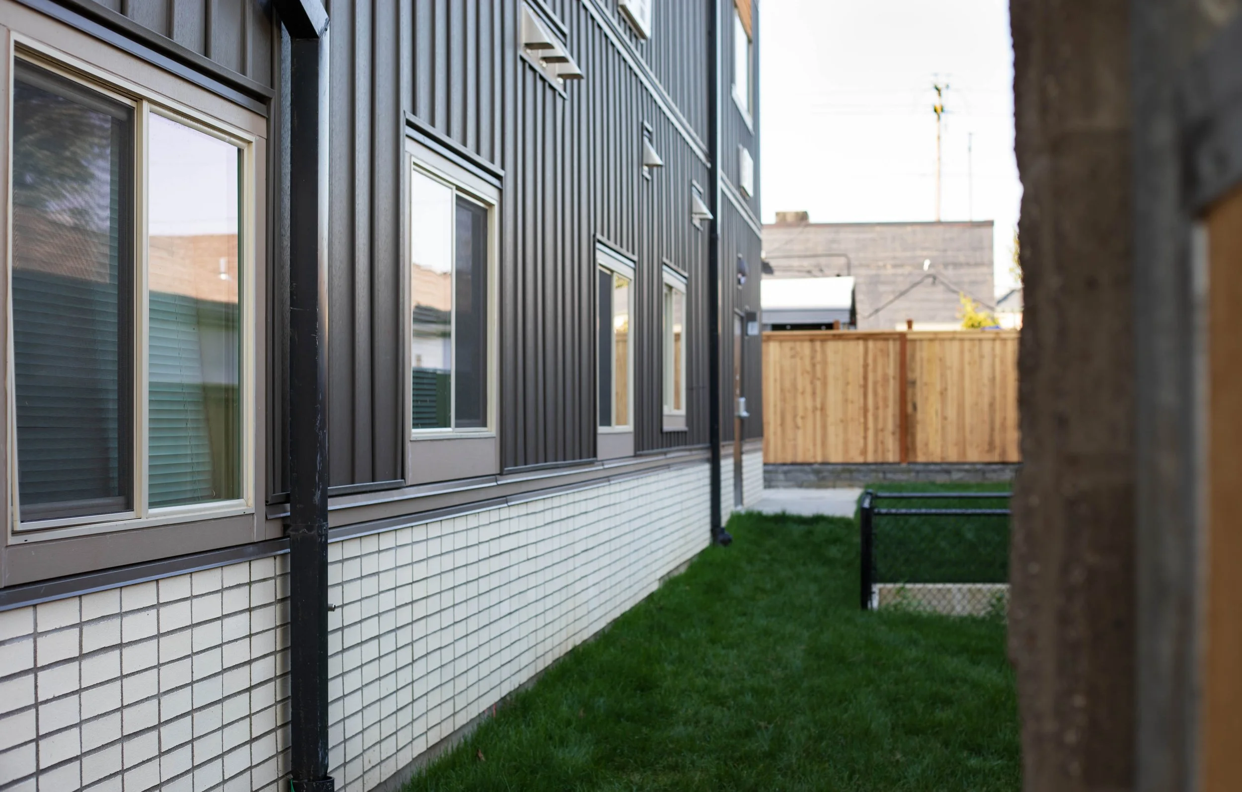 Side of a modern residential building with black metal siding, multiple windows, a white tiled lower facade, and a green lawn yard enclosed by a wooden fence.