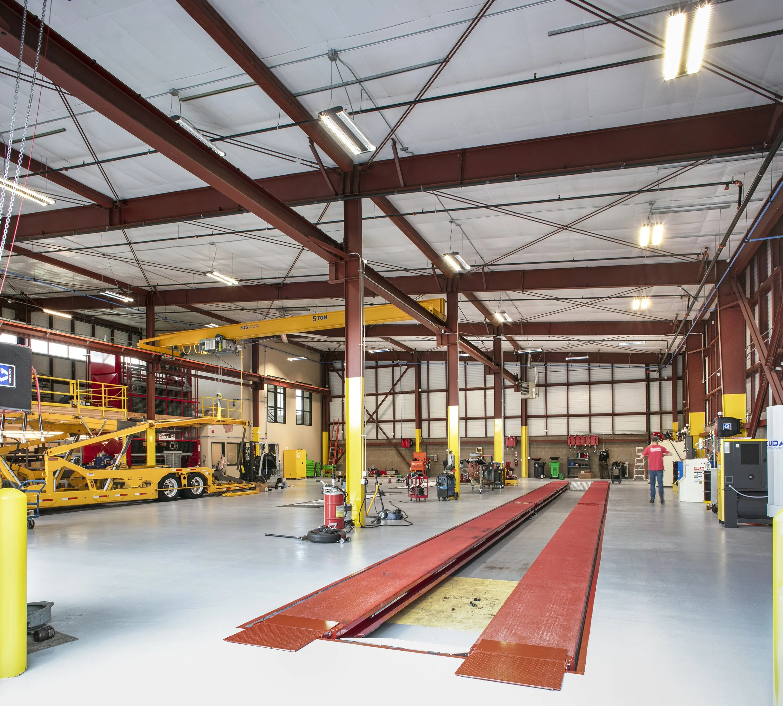 A spacious industrial workshop with yellow and red hydraulic lifts, a yellow overhead crane, workbenches, and tools. The workshop has high ceilings with bright lighting and a gray floor.