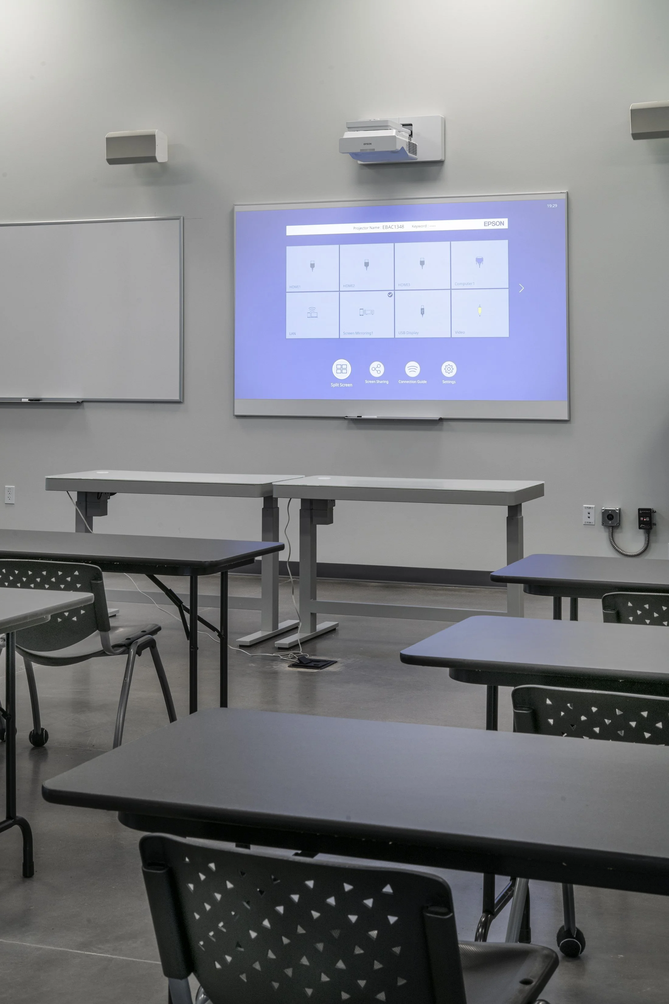 Classroom with desks, a whiteboard, and a digital projector screen displaying a setup menu, with a wall-mounted whiteboard and electrical outlets visible.