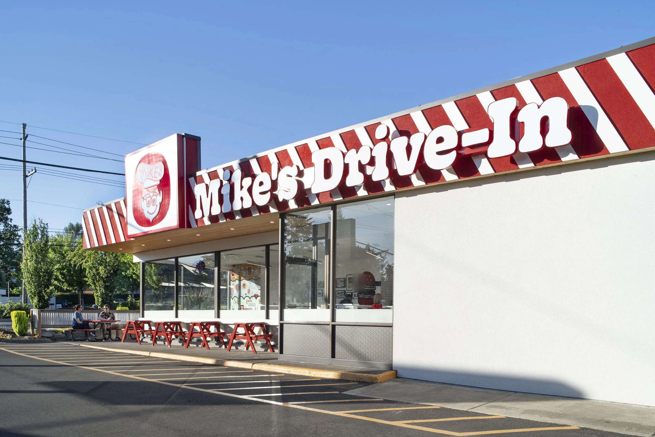 Exterior of Mike's Drive-In restaurant with red and white striped sign, outdoor seating, and a mostly clear blue sky.