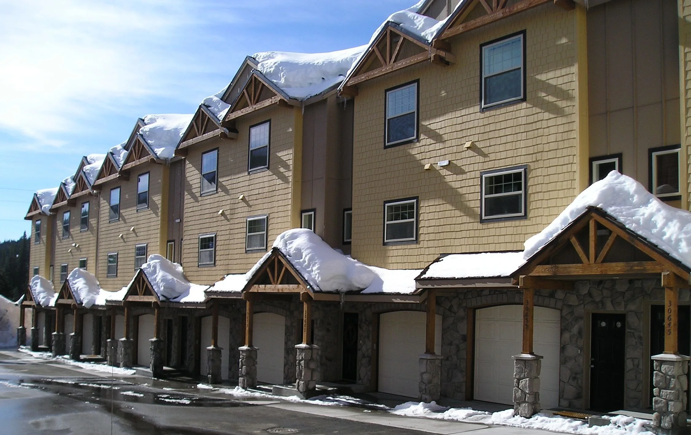 Multi-story residential building with tan siding, stone and wood porch supports, and snow-covered roofs, with snow on the ground and a clear blue sky.