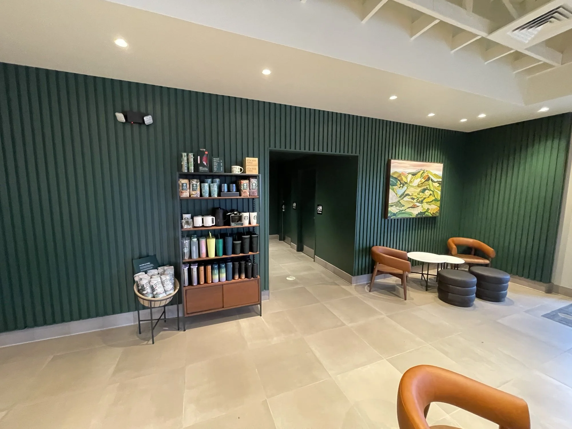 Indoor seating area with green textured wall, a colorful landscape painting, a small coffee table, and two brown chairs with round black ottomans. A shelf displays coffee mugs and reusable cups. A small basket holds coffee packets, and ceiling lights