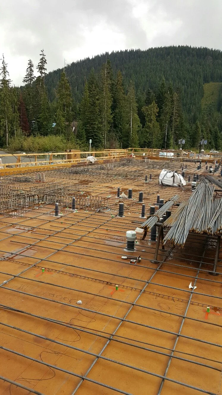 Construction site for a building with reinforcing steel rebar and formwork, surrounded by a rope safety barrier, with a forested mountain and cloudy sky in the background.