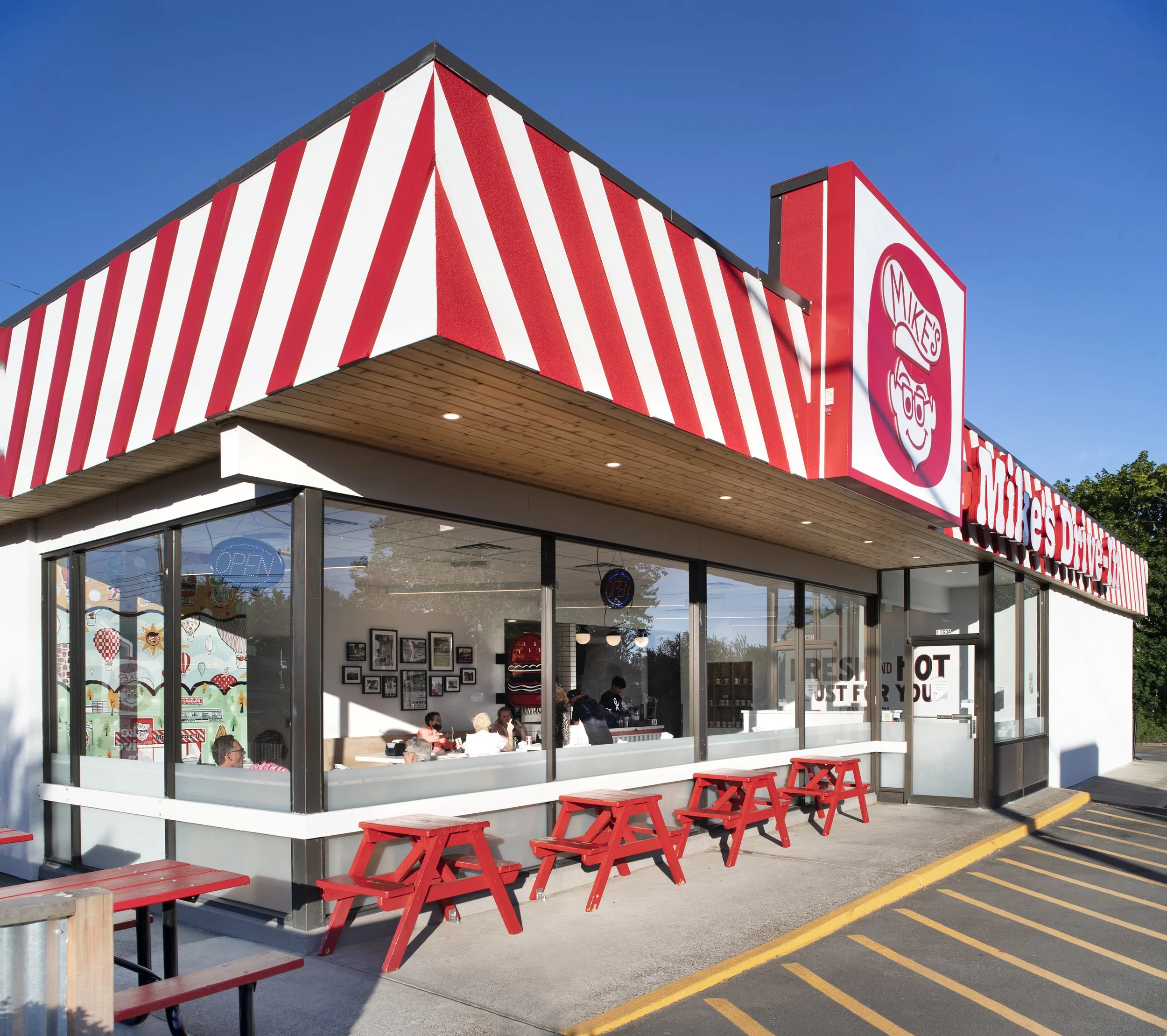 Exterior of a Clyde's restaurant with red and white striped awning, glass windows showing customers inside, red picnic tables outside, and a large sign with a burger and ice cream logo.