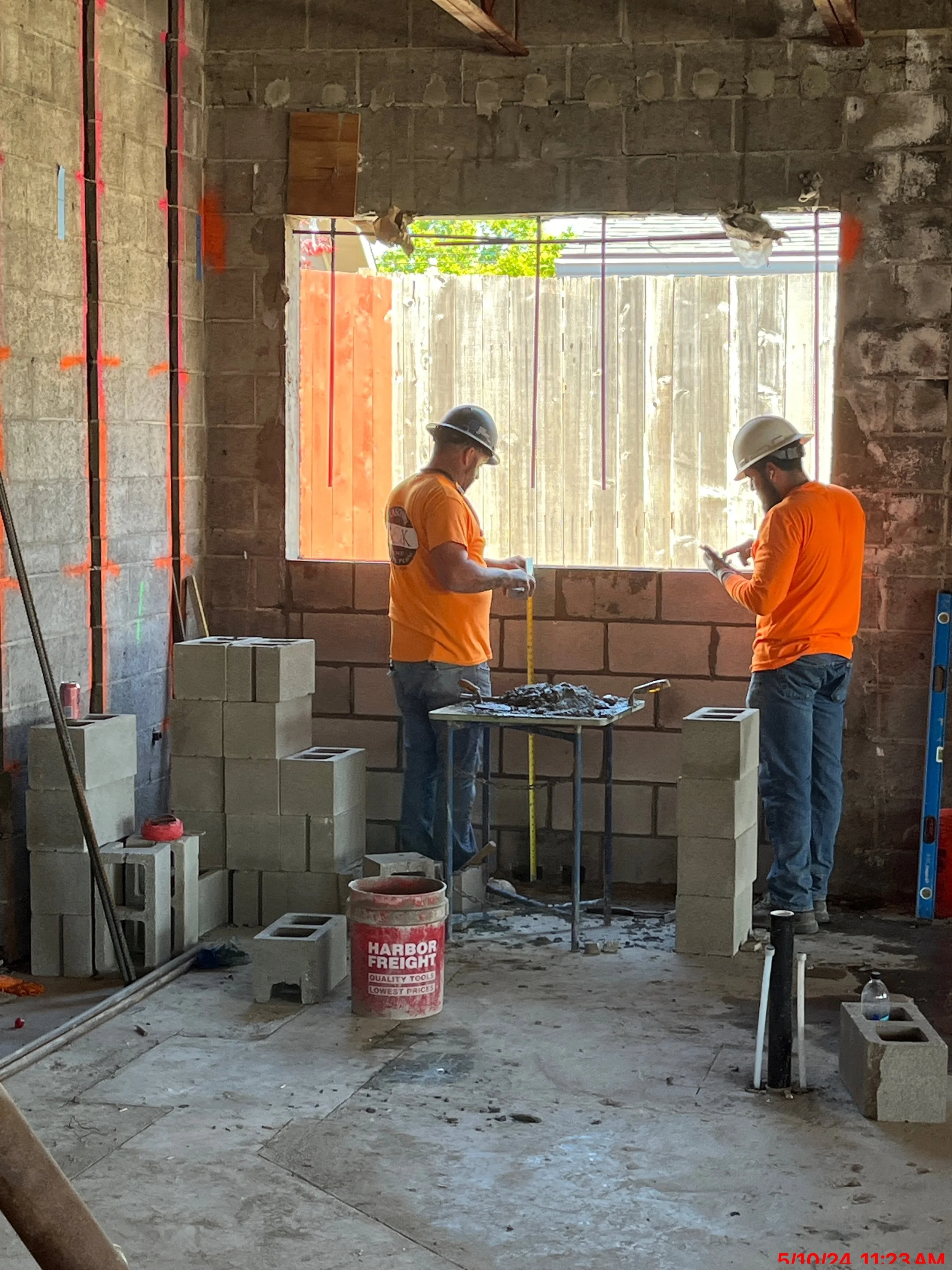Two construction workers in orange shirts and white helmets working inside a building under construction, with cinder blocks and tools around, and a large open window/opening in the wall.