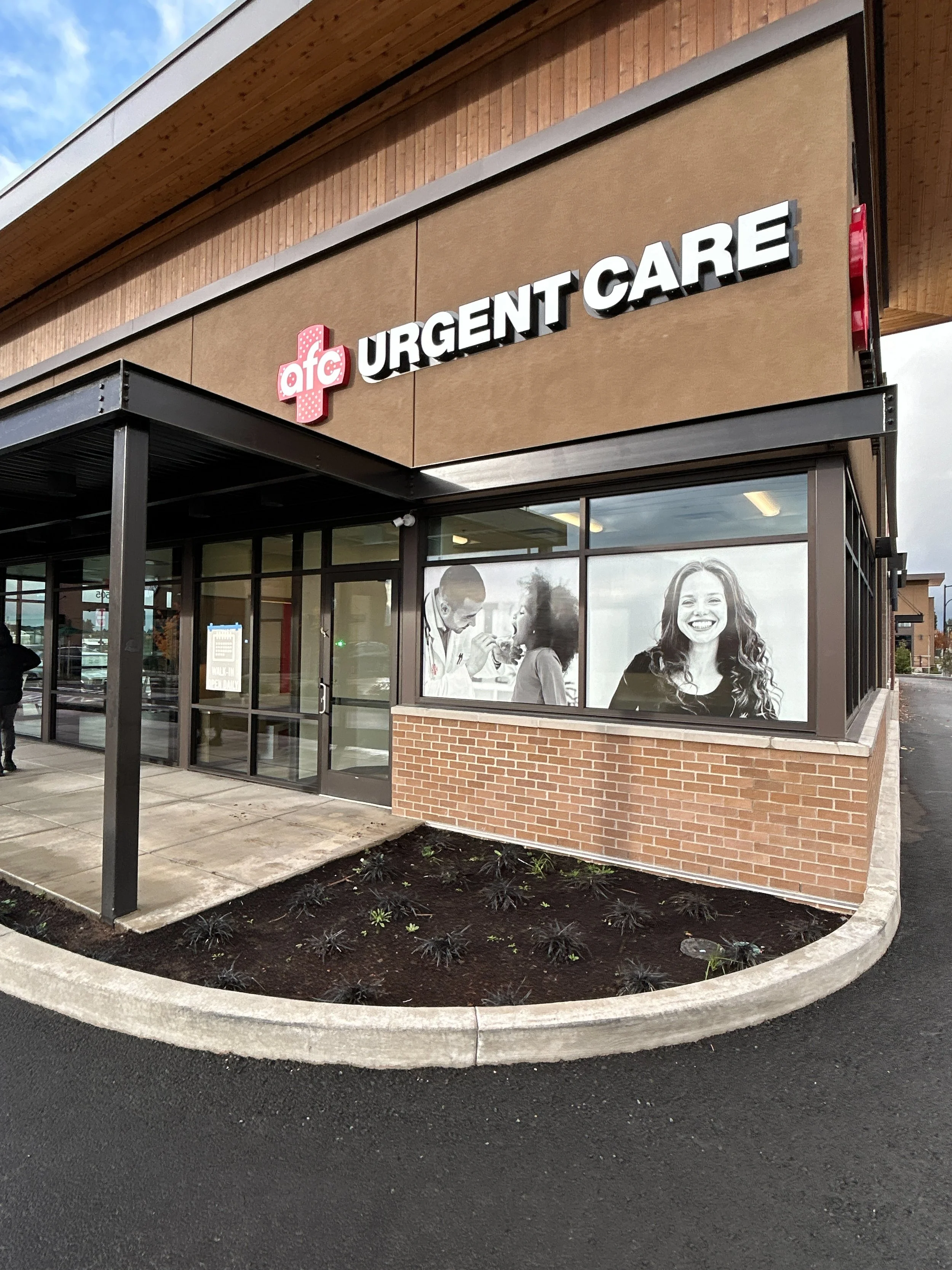 Exterior of an AFC Urgent Care clinic with a brown and brick facade, glass windows, and a large smiley face poster in the window.