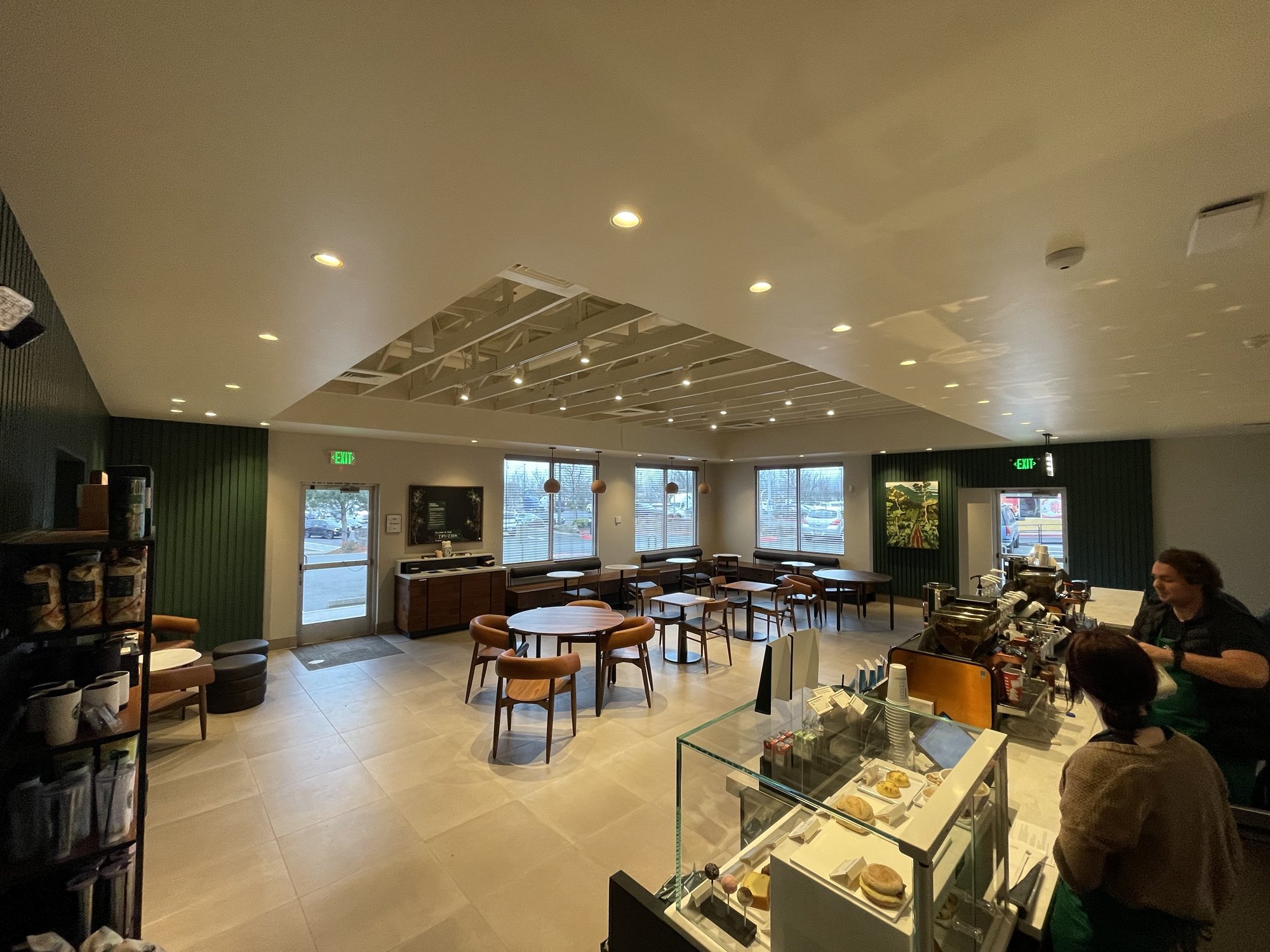 Interior of a modern coffee shop with seating, a display case of baked goods, an espresso bar, and two staff members working behind the counter.