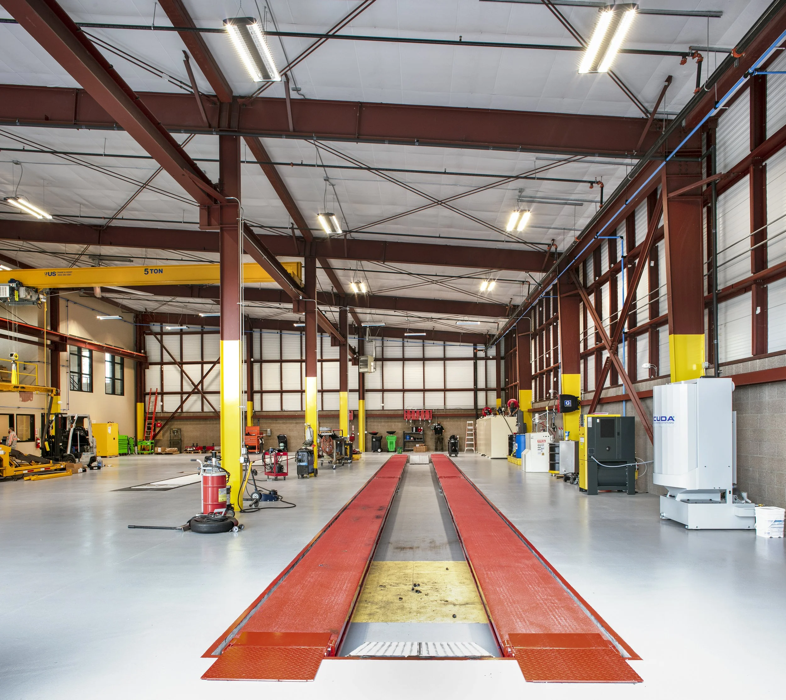A spacious industrial workshop with red metal beams, yellow support posts, and a central red and yellow vehicle lift. There are various tools, equipment, and machinery along the walls, with a high ceiling and large windows letting in natural light.