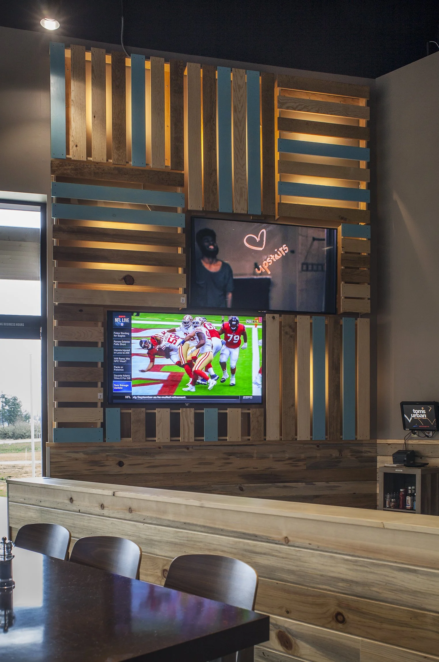 Interior of a modern room with a wooden slat wall and two mounted flat-screen TVs showing a football game and a neon sign, with a lounge seating area and a gathering table in the foreground.