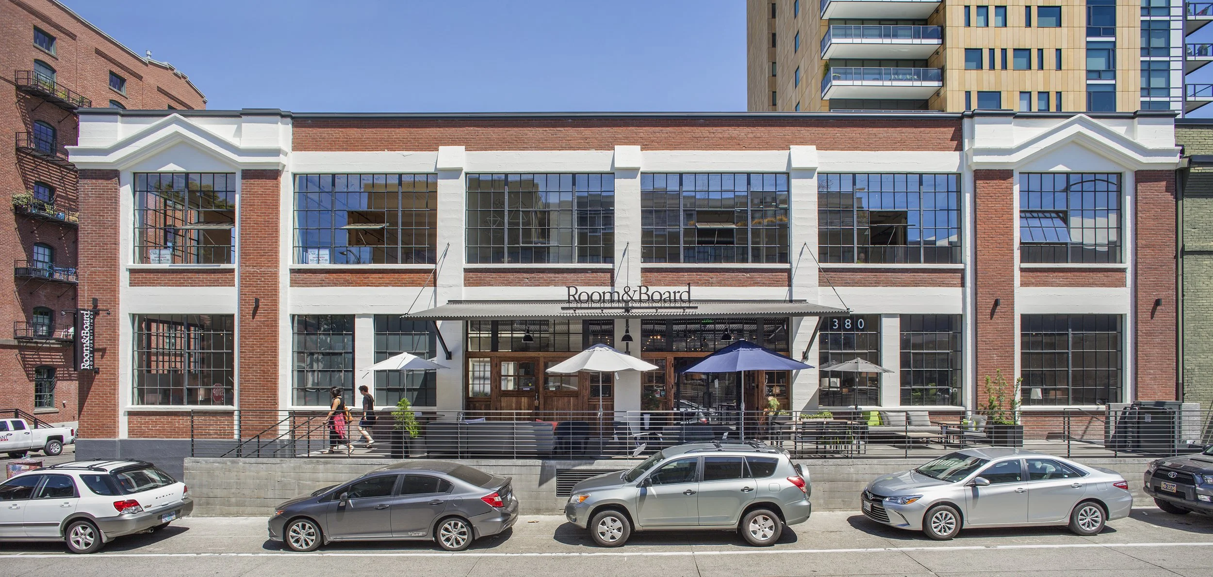 Exterior of a modern brick building with large windows and a sign that reads 'Room & Board.' There is an outdoor seating area with umbrellas and a railing, and cars parked in front.