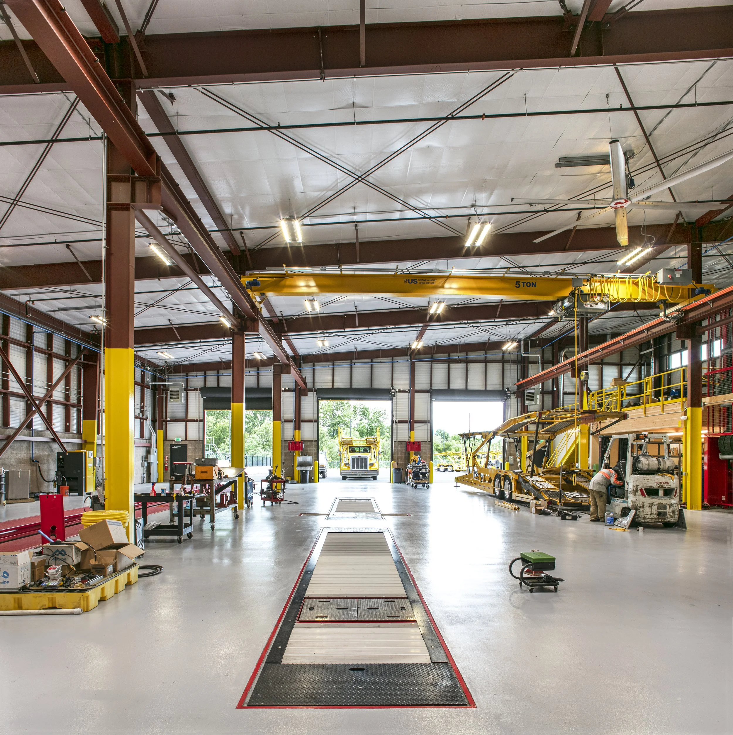 An industrial workshop with a high ceiling, large overhead crane, and various machinery and equipment, including trucks and a forklift, in a spacious, well-lit environment.