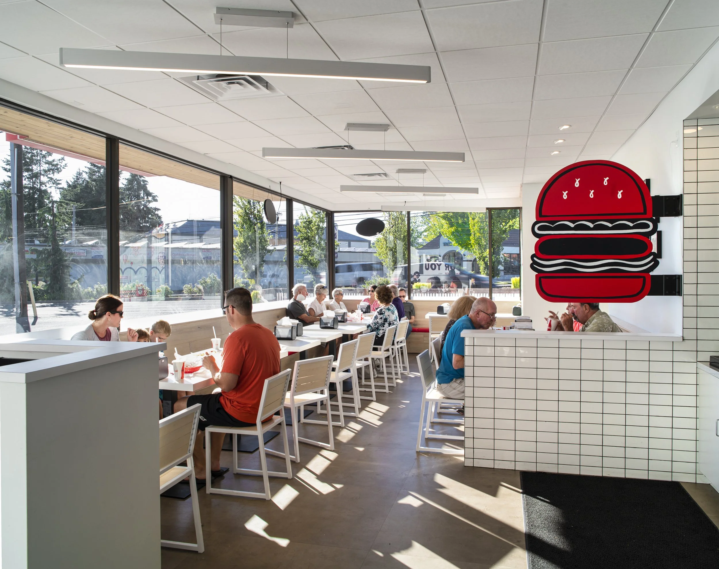Interior of a modern burger restaurant with large windows, white tiled walls, and a bright atmosphere. There are several customers seated at white tables, enjoying their meals.