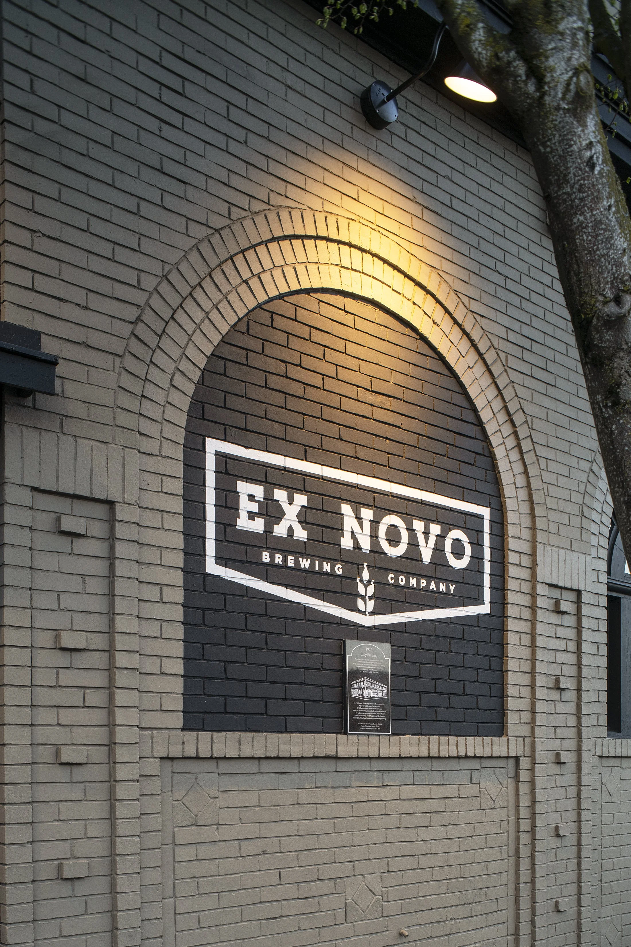 Sign on a brick wall for EX NOVO Brewing Company, with a tree branch overhead and a light illuminating the sign.