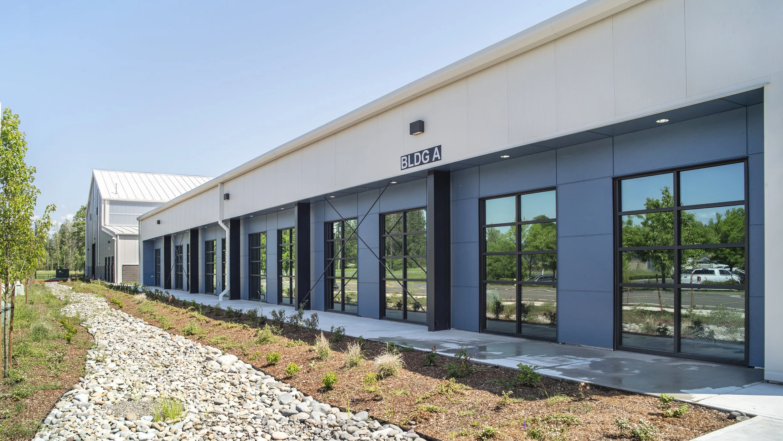 Exterior view of a modern commercial building labeled BLDG A with large glass windows, landscaped with plants and rocks, under a clear blue sky.