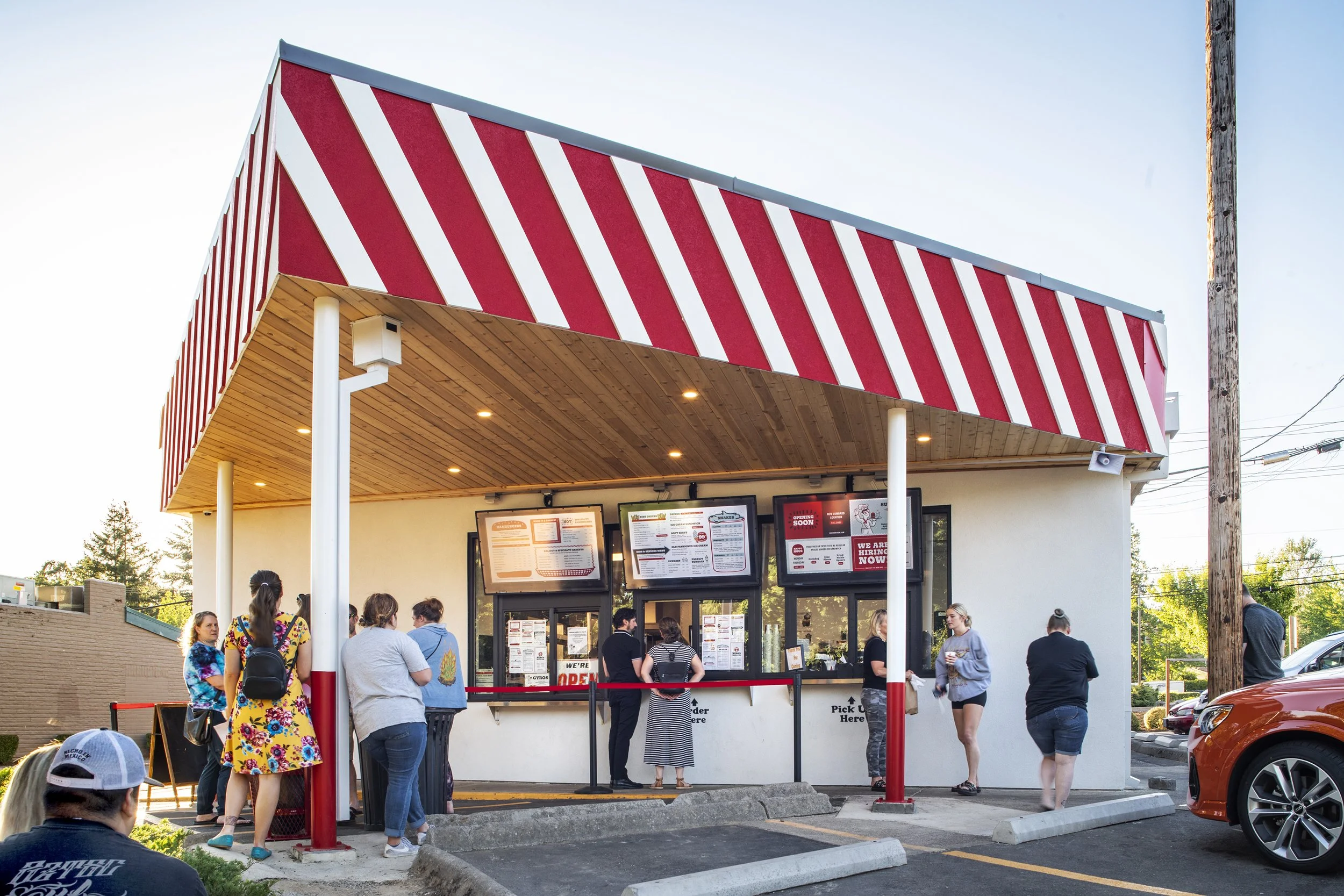 People standing in line at a fast-food restaurant window with digital menu boards, in a parking lot next to a building with a red and white striped awning.