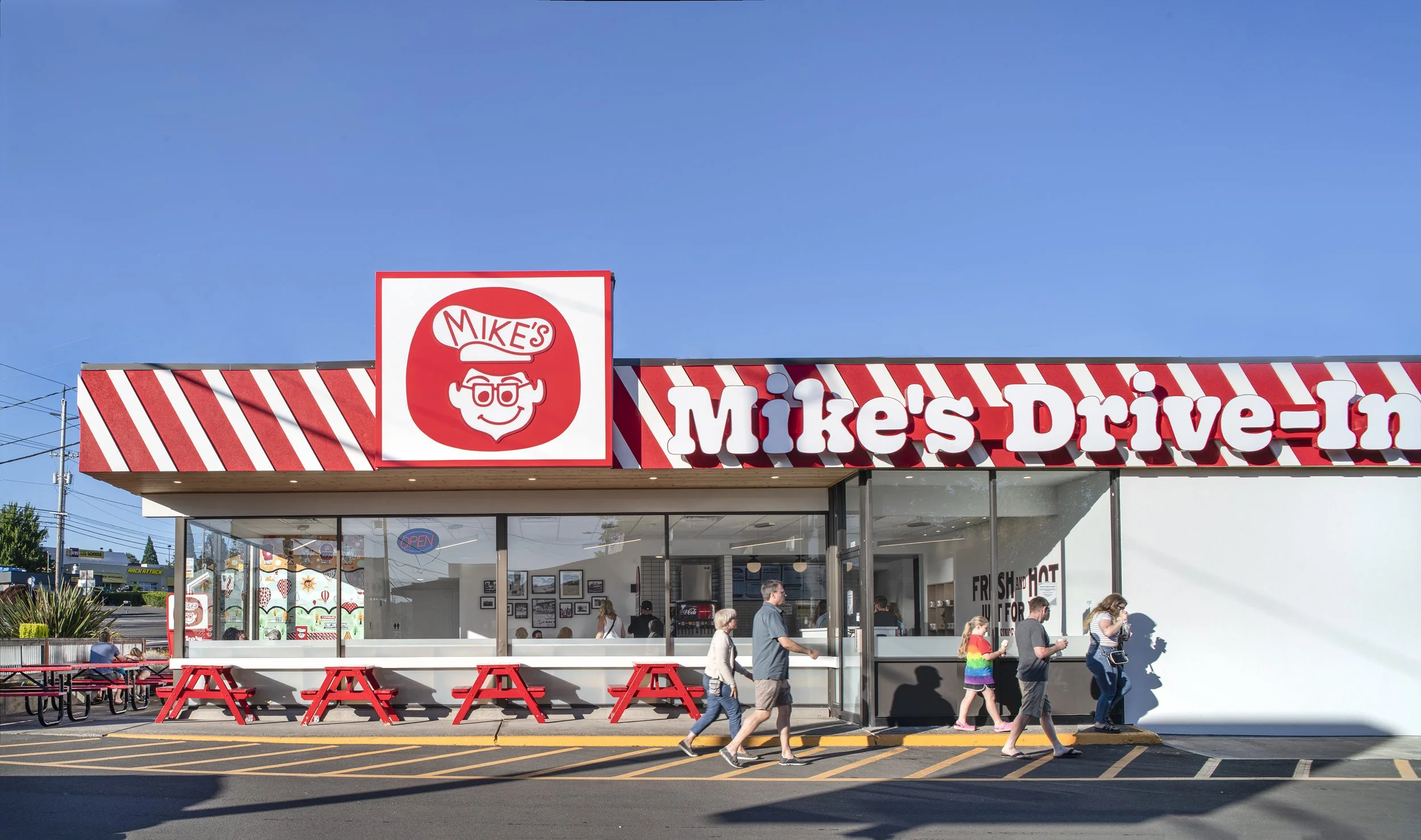 Front of Mike's Drive-In, a restaurant with a red and white striped exterior, large sign with a cartoon face and glasses, and people walking by during daytime.